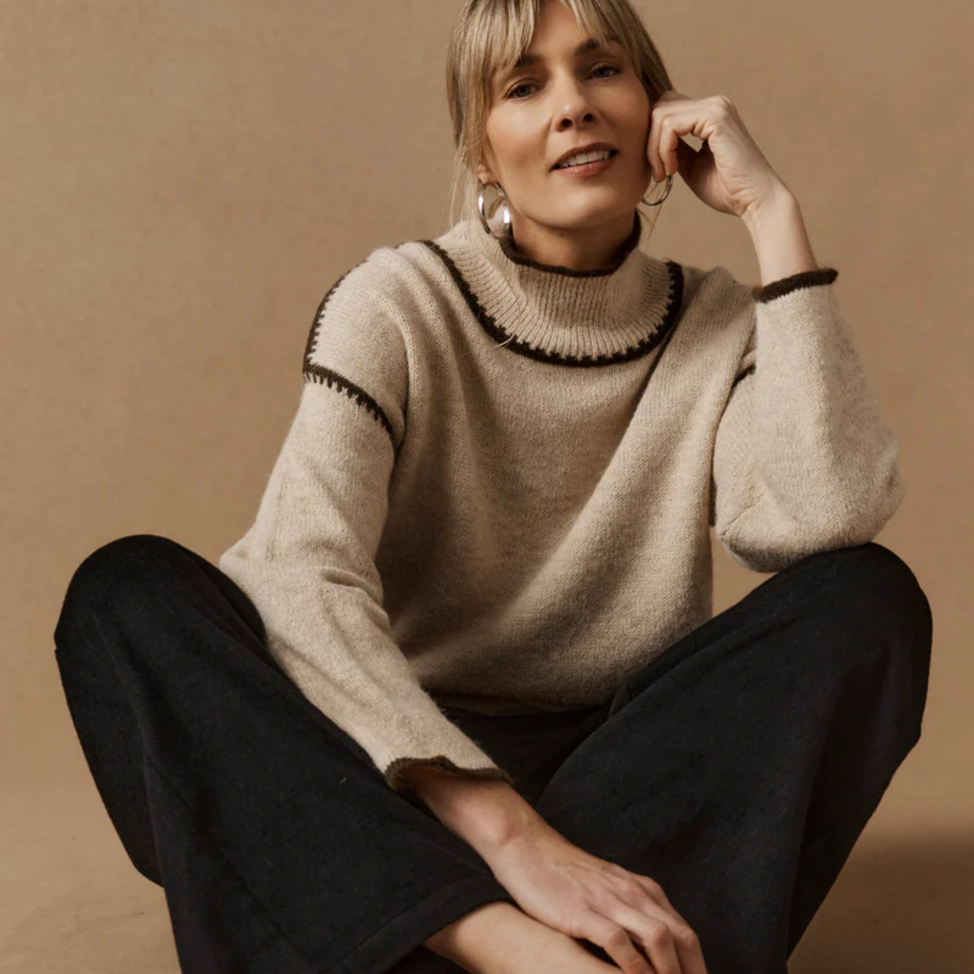 A woman with short blonde hair and hoop earrings is sitting cross-legged against a beige background, wearing a beige sweater with black trim, with her hand resting on her face.