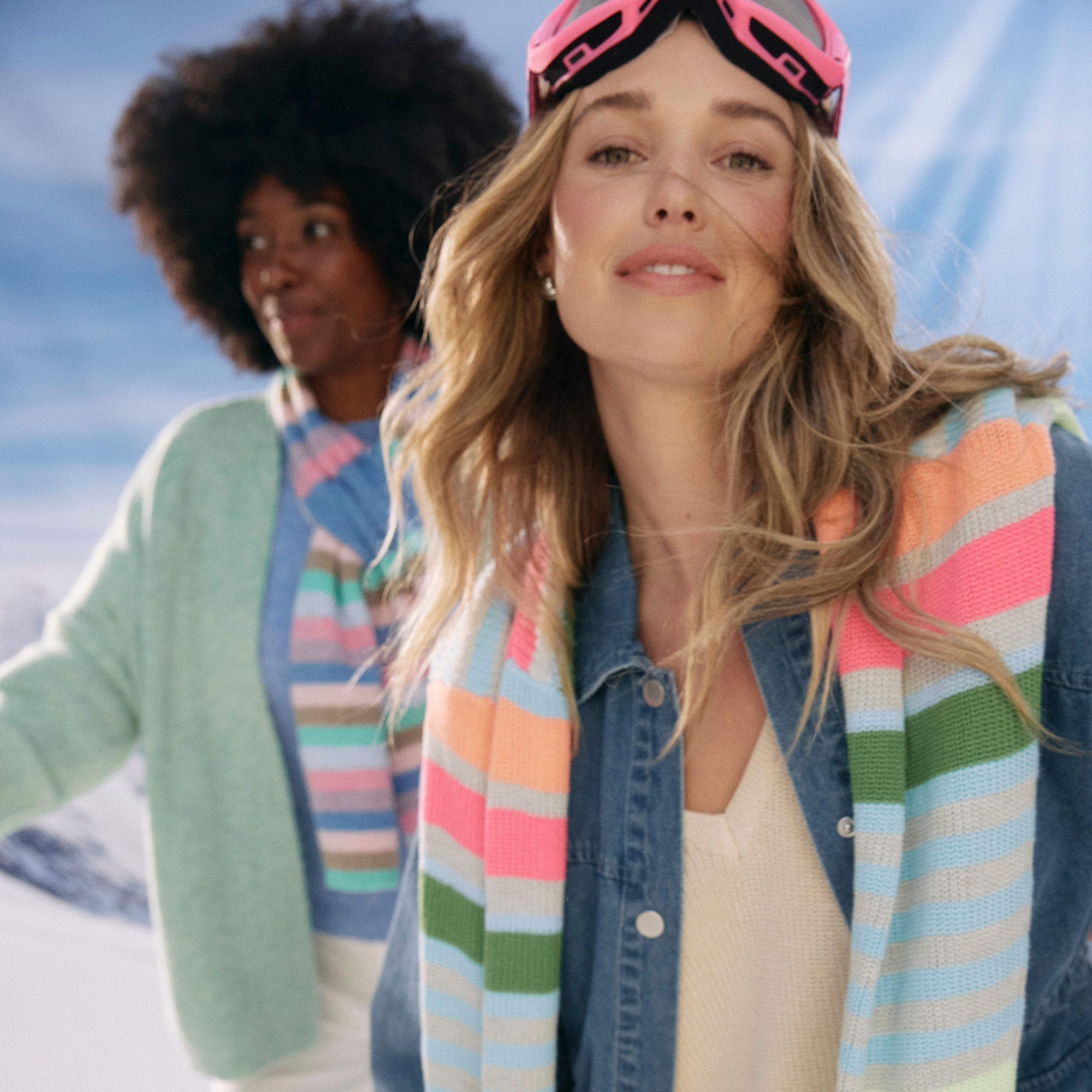 Close-up of a woman with long blonde hair wearing pink ski goggles and a colorful striped sweater, smiling with a partly cloudy sky in the background.