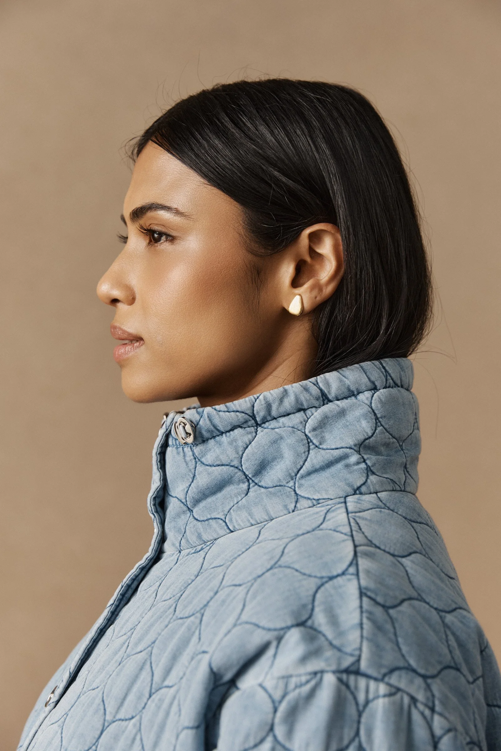 Close-up profile of a woman with dark, straight hair, wearing a quilted, light blue jacket and gold earrings, against a beige background.