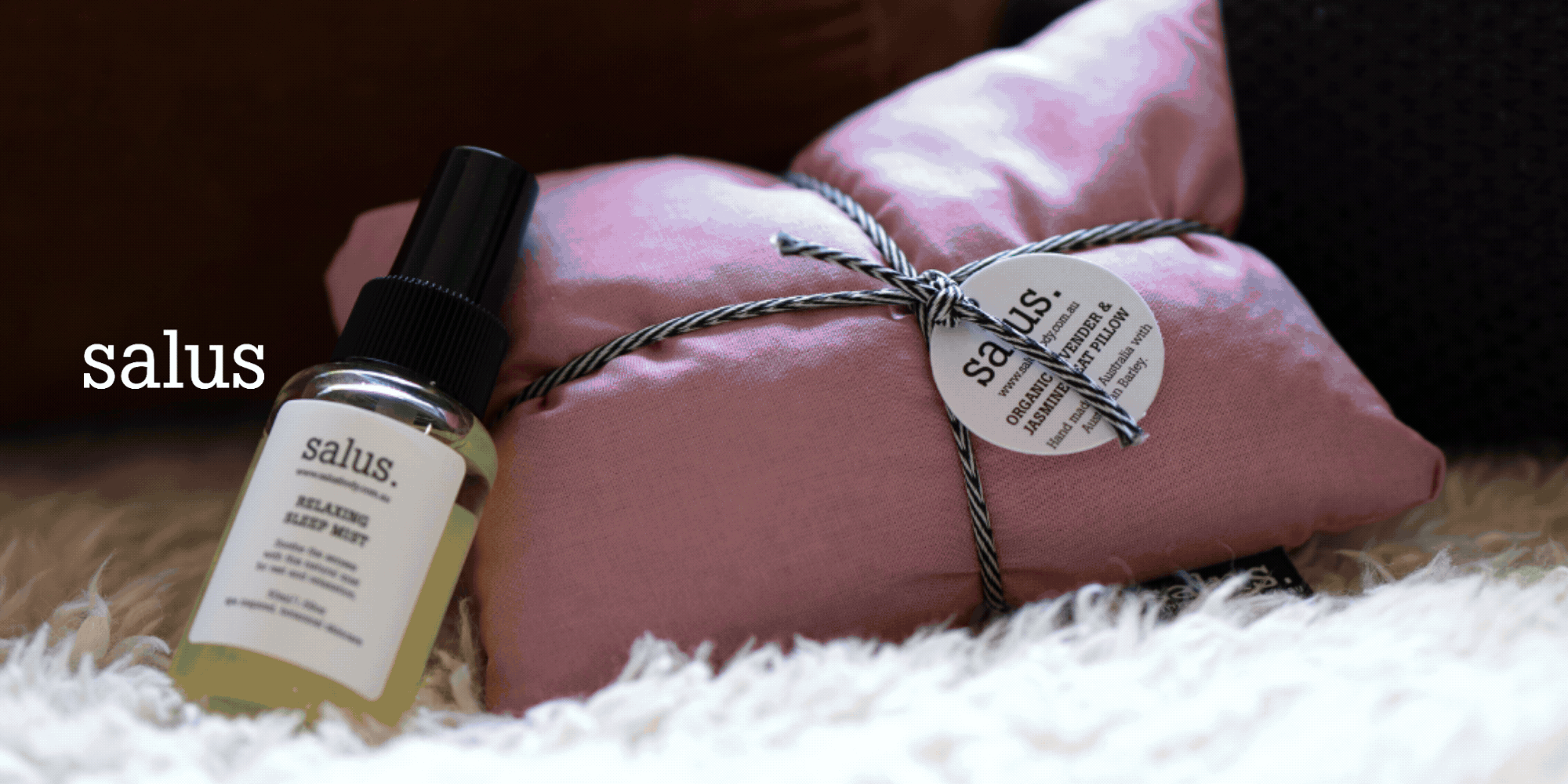 A small bottle of salus relaxing herbal mist and a pink wrapped gift tied with black and white striped twine, with a salus tag, resting on a white fluffy surface.