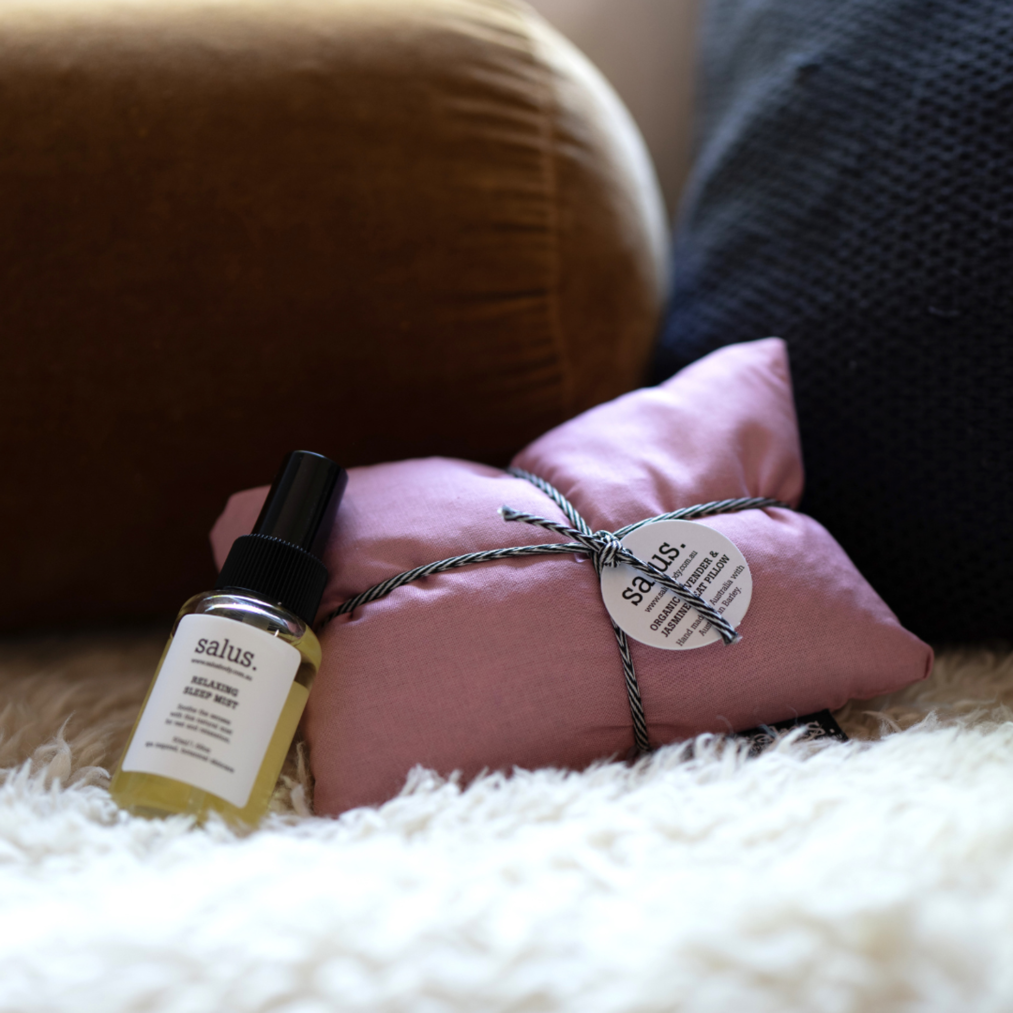 A pink heat pack pillow with a black and white striped cord tied around it, a tag with the brand name 'salus' is partly visible, in front of a golden-brown pillow, placed underneath a white fluffy blanket on the floor.
