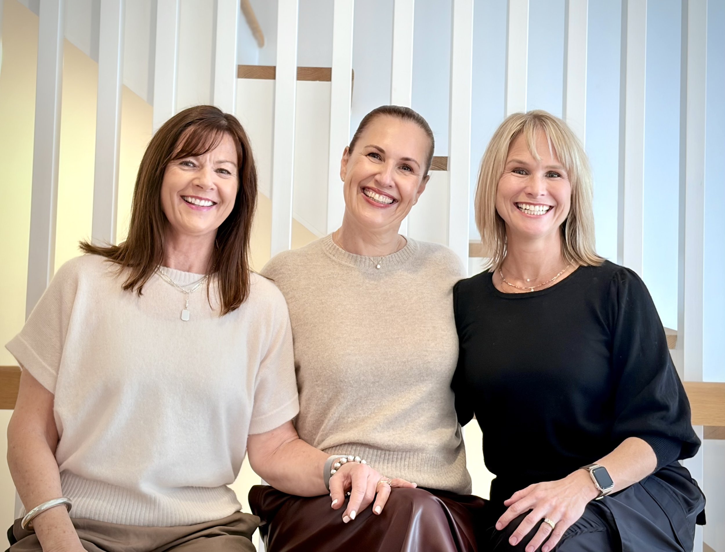 Three women with big smiles posing together in an indoor setting with a modern staircase in the background, wearing casual tops and jewelry.