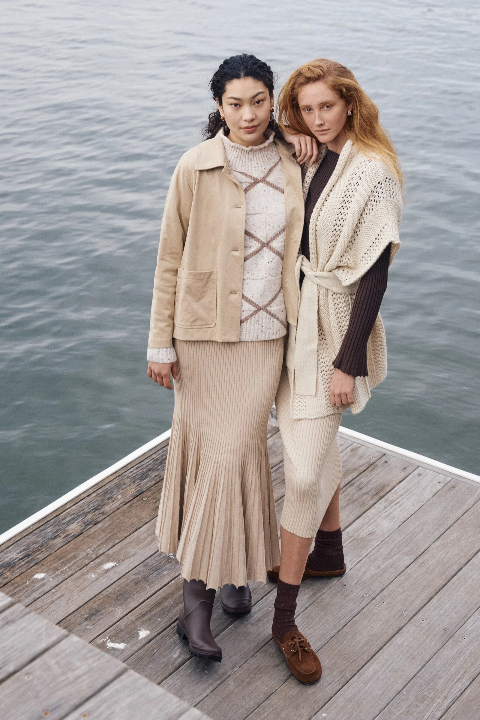 Two women standing on a wooden dock by a body of water, posing for a photo. One woman has black hair and is wearing a beige coat, patterned sweater, and long pleated skirt; the other has red hair and is dressed in a cream knitted vest, dark top, and 