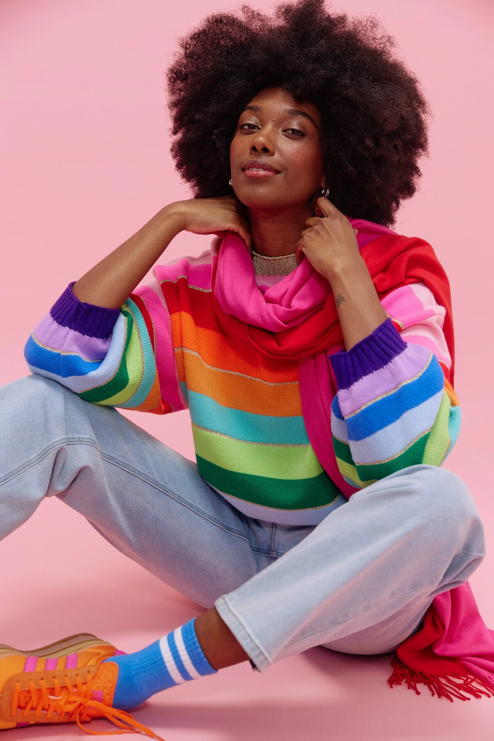 A woman with dark skin and natural, afro hairstyle posing against a pink background. She is wearing a colorful, rainbow-striped sweater, a pink scarf, light blue jeans, and orange sneakers with blue striped socks. She has silver earrings and a small 