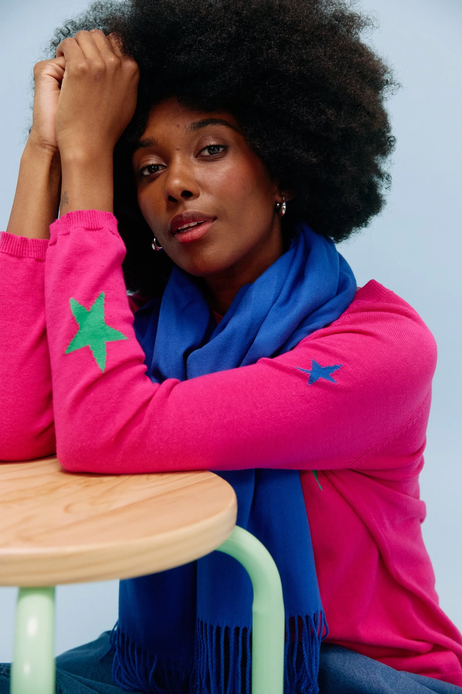 A young woman with dark skin and curly black hair sitting next to a light-colored wooden table with pastel green legs, wearing a pink sweater with colorful star patches, a blue scarf, and dark blue pants with fringes.