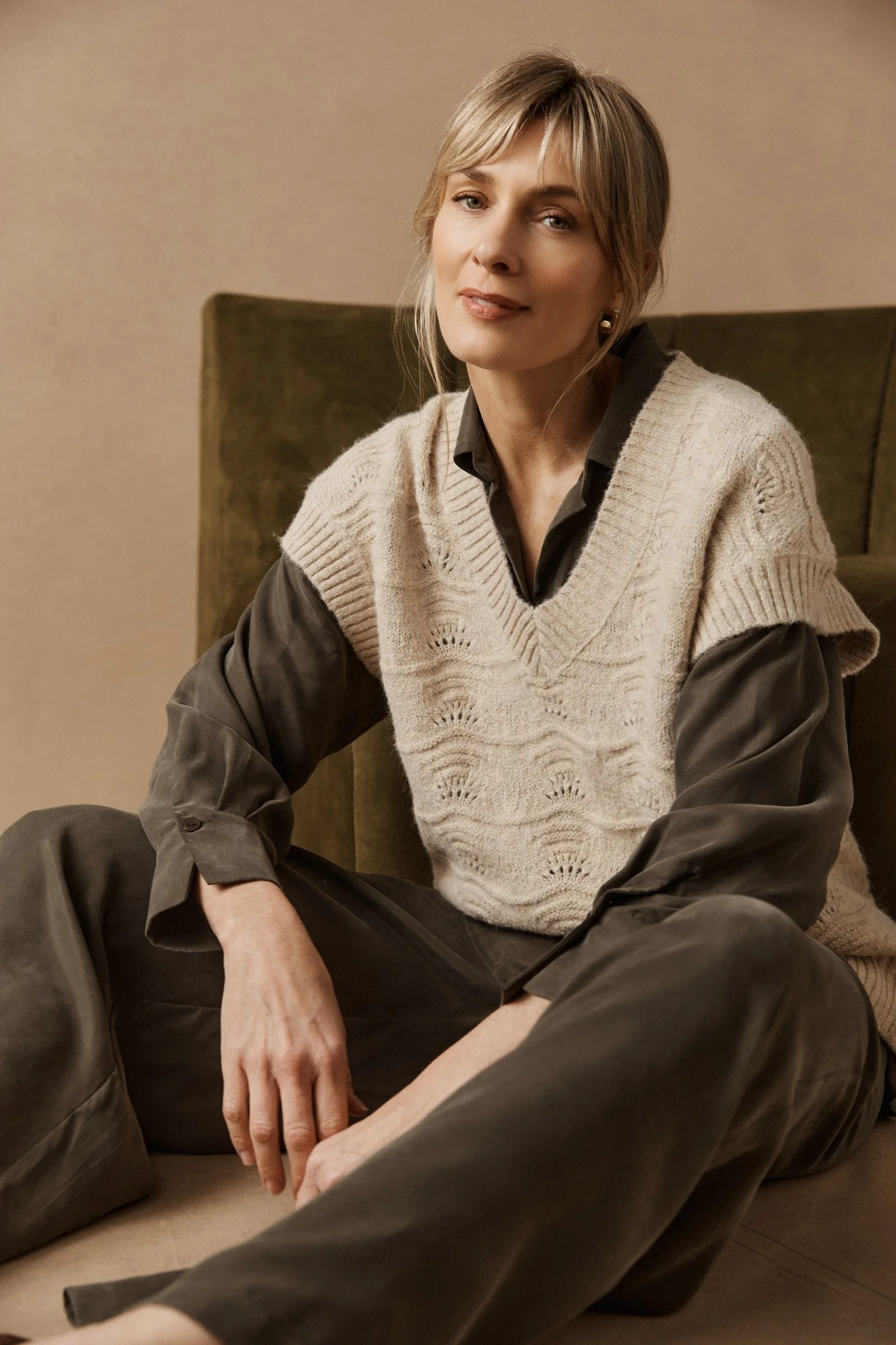 A woman with blonde hair, wearing a beige knit vest over a black shirt, sitting on the floor against a beige background. She is looking at the camera with a slight smile.