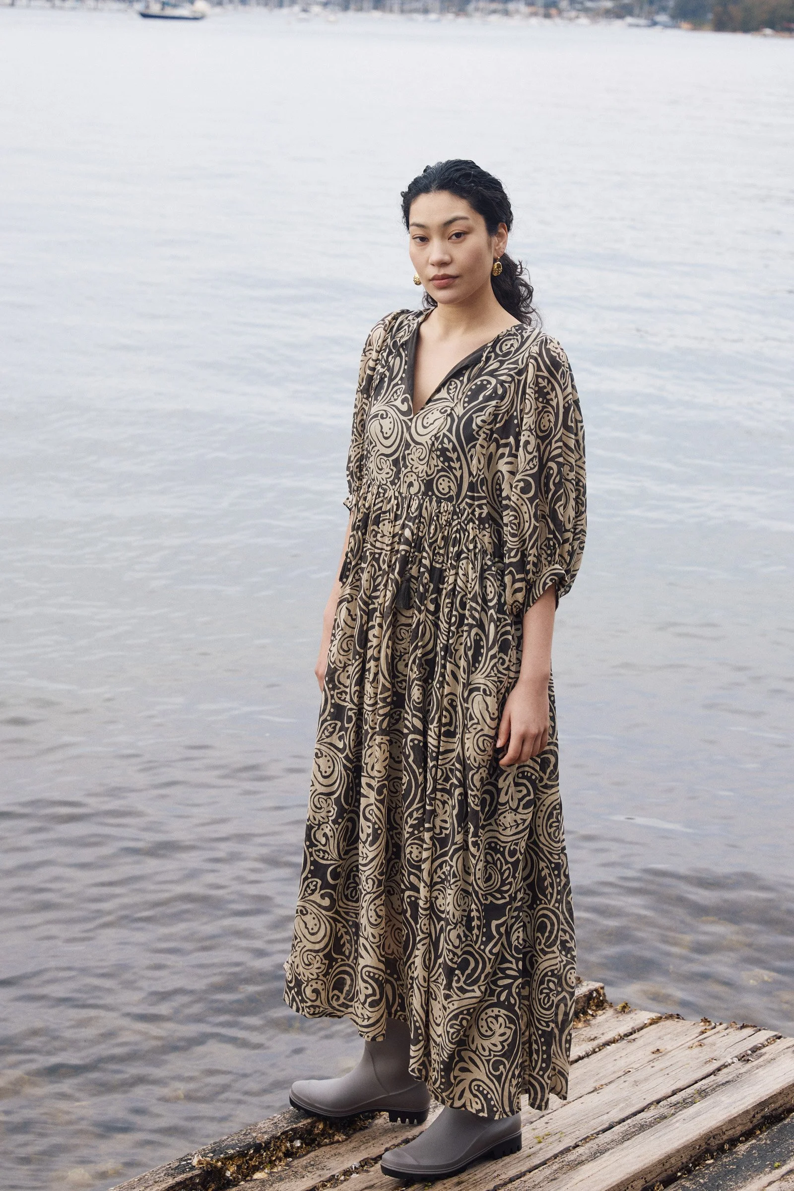 A woman standing on a wooden dock by the water, wearing a long patterned dress, gray rain boots, gold earrings, and with dark curly hair, during a cloudy day.