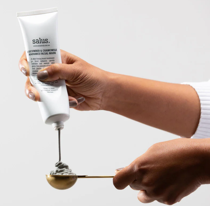 Hand squeezing cucumber and chamomile facial mask onto a gold spoon. Salus body products