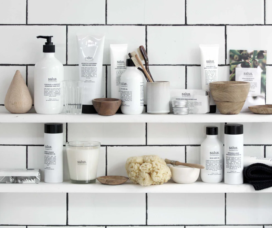 Bathroom shelves with skincare from Salus body, haircare products, a candle, a sponge, and natural wooden bowls against a white tiled wall.