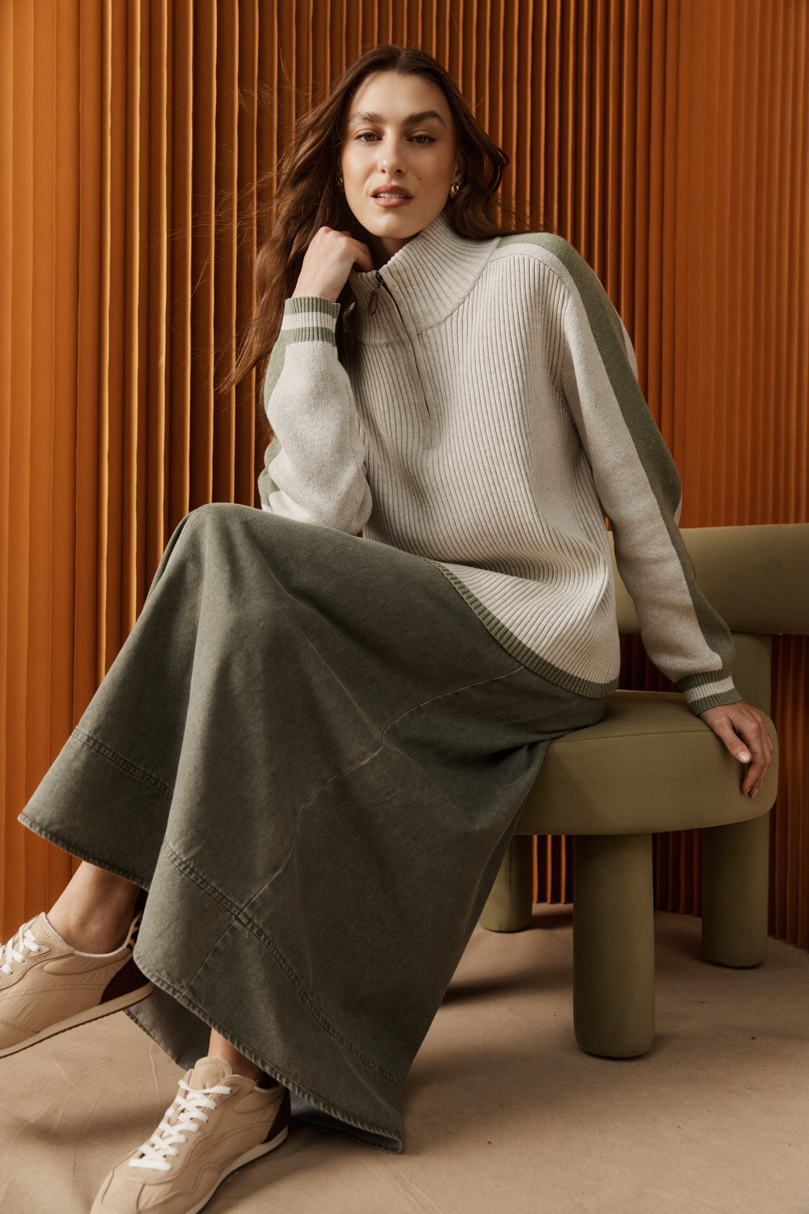 A woman with long brown hair sitting on a beige chair, wearing a cream turtleneck sweater with green accents, a long dark skirt, and beige sneakers. She is posing with her left hand on her chin against a background of vertical wooden slats.