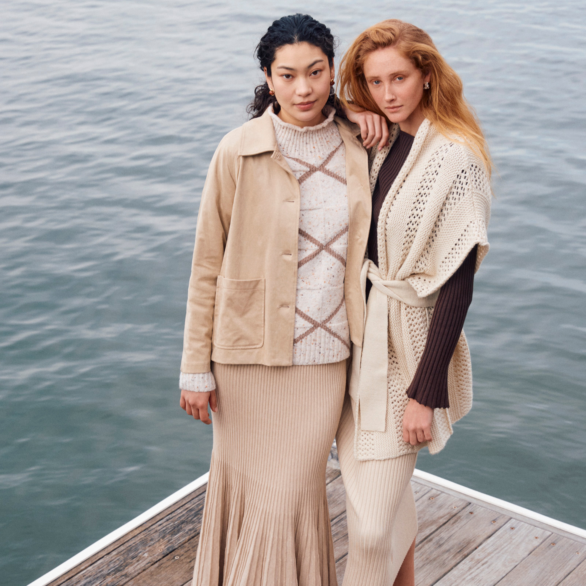 Two women standing on a dock by the water, wearing beige and cream-colored knit and wool clothing, with one woman having black hair and the other red hair.