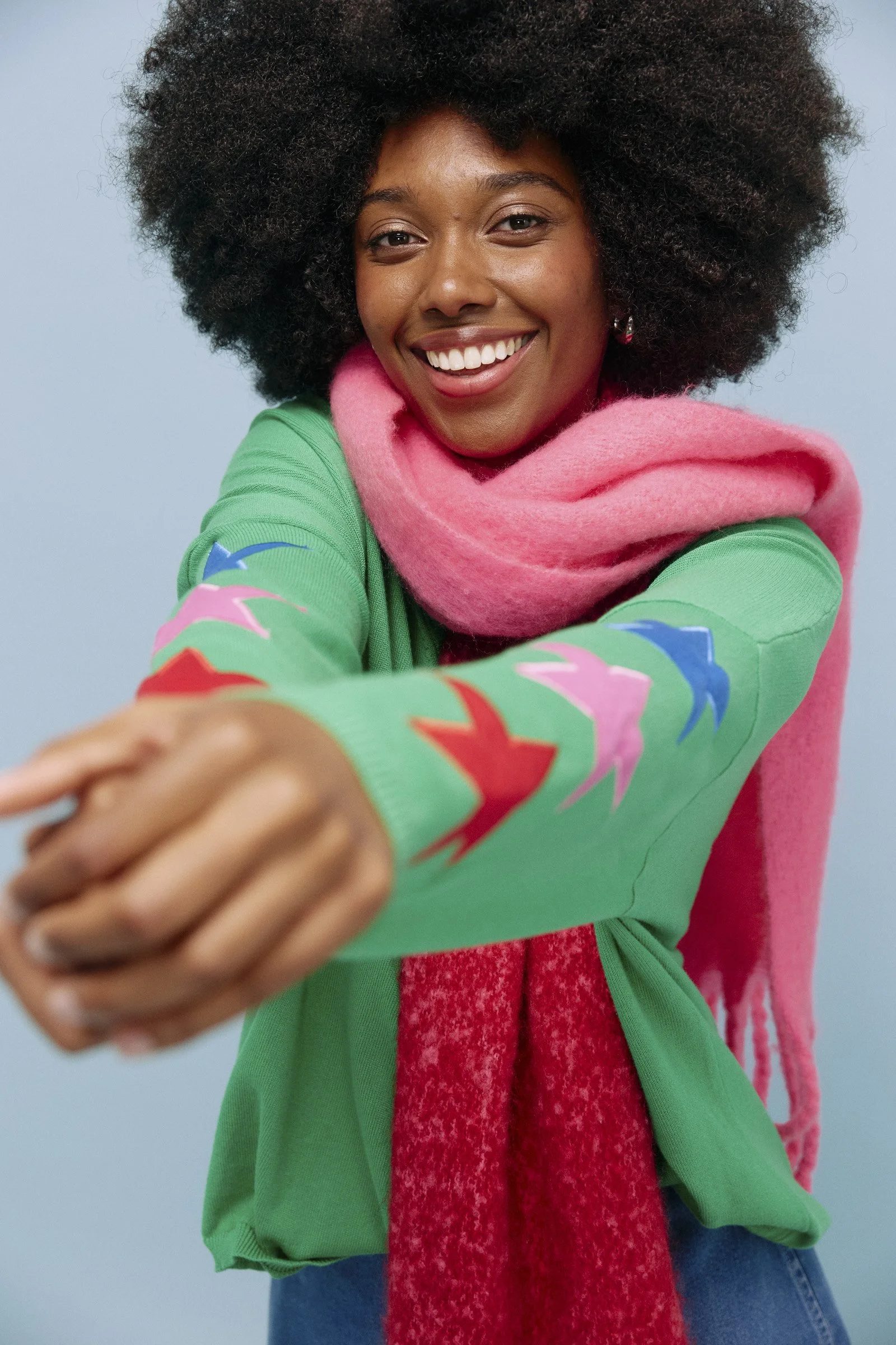 A young woman with dark curly hair smiling and reaching toward the camera, wearing a green sweater with colorful patterns on the sleeves, a pink scarf, and blue jeans against a light blue background.
