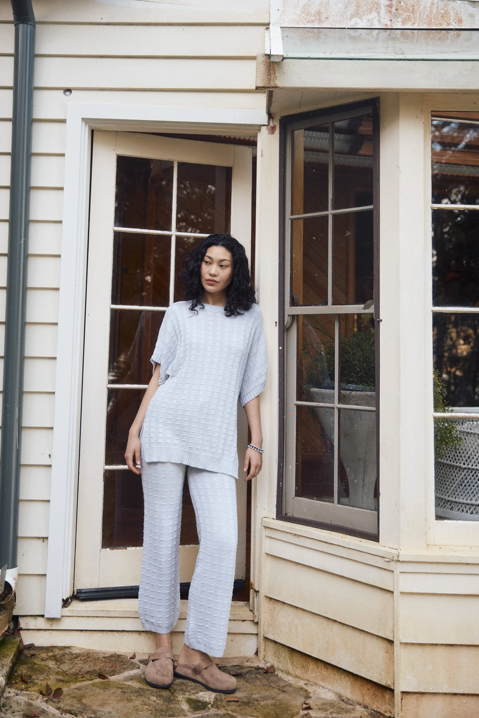 Woman with curly black hair wearing a loose gray checkered sweater and matching pants standing outside next to house with cream-colored siding and two windows, one open, on a stone pathway.