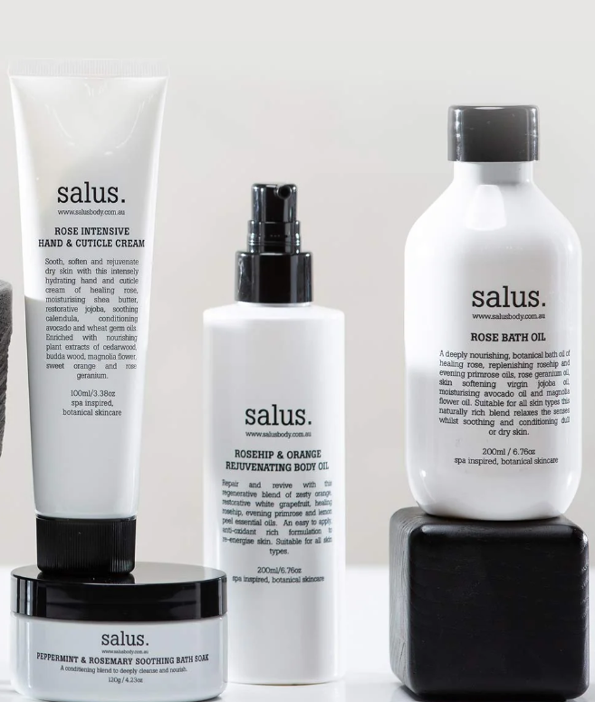 Collection of skincare products including a tube of Salus hand and cuticle cream, a bottle of Salus rosehip and orange body oil, a bottle of Salus rose bath oil, and a jar of Salus peppermint and rosemary soothing bath soak, arranged on black and whi