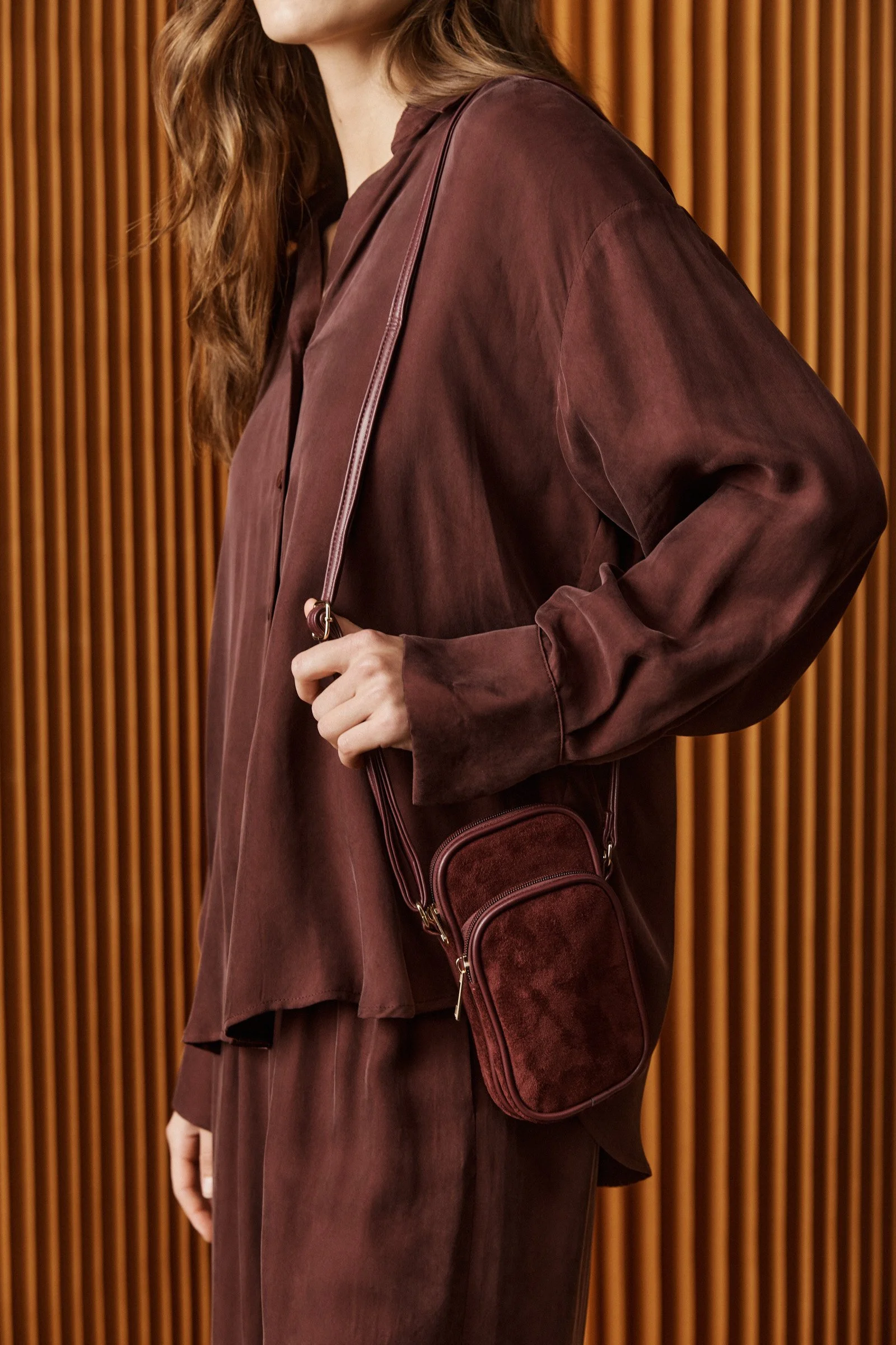 A woman is wearing a long, loose, burgundy shirt with matching pants, holding a small burgundy crossbody bag, standing in front of a wooden panel background.