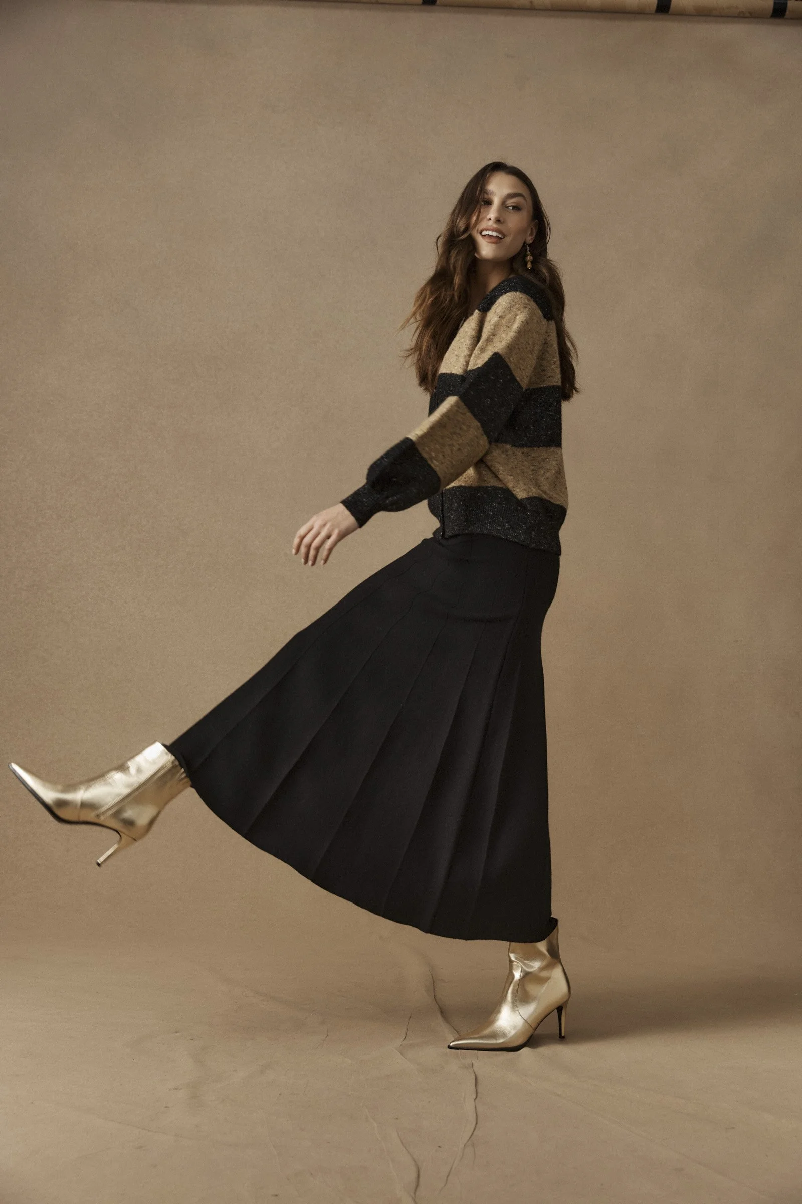 Woman with long brown hair wearing metallic gold high heel boots, a long black pleated skirt, and a beige and black oversized sweater, posing against a plain beige background.