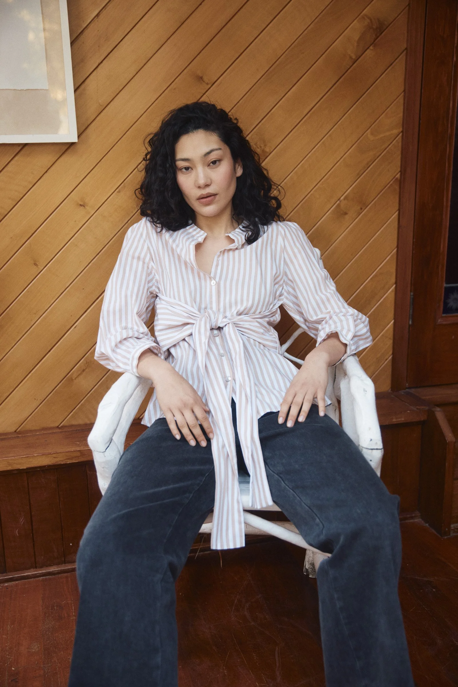 A woman with curly black hair sitting in a white chair against a wooden wall, wearing a striped shirt tied at the waist and black pants.