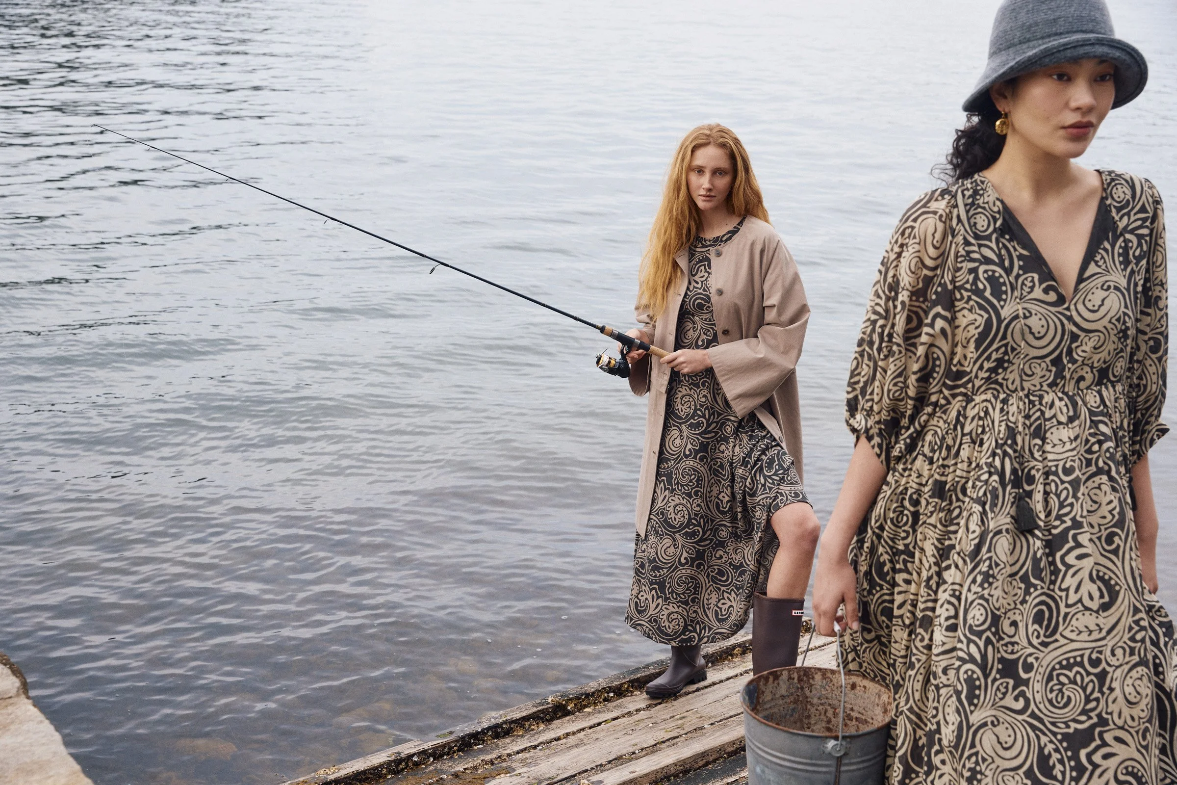 Two women standing on a dock by the water, one fishing with a rod and the other in a patterned dress and hat, with a rusty bucket nearby.