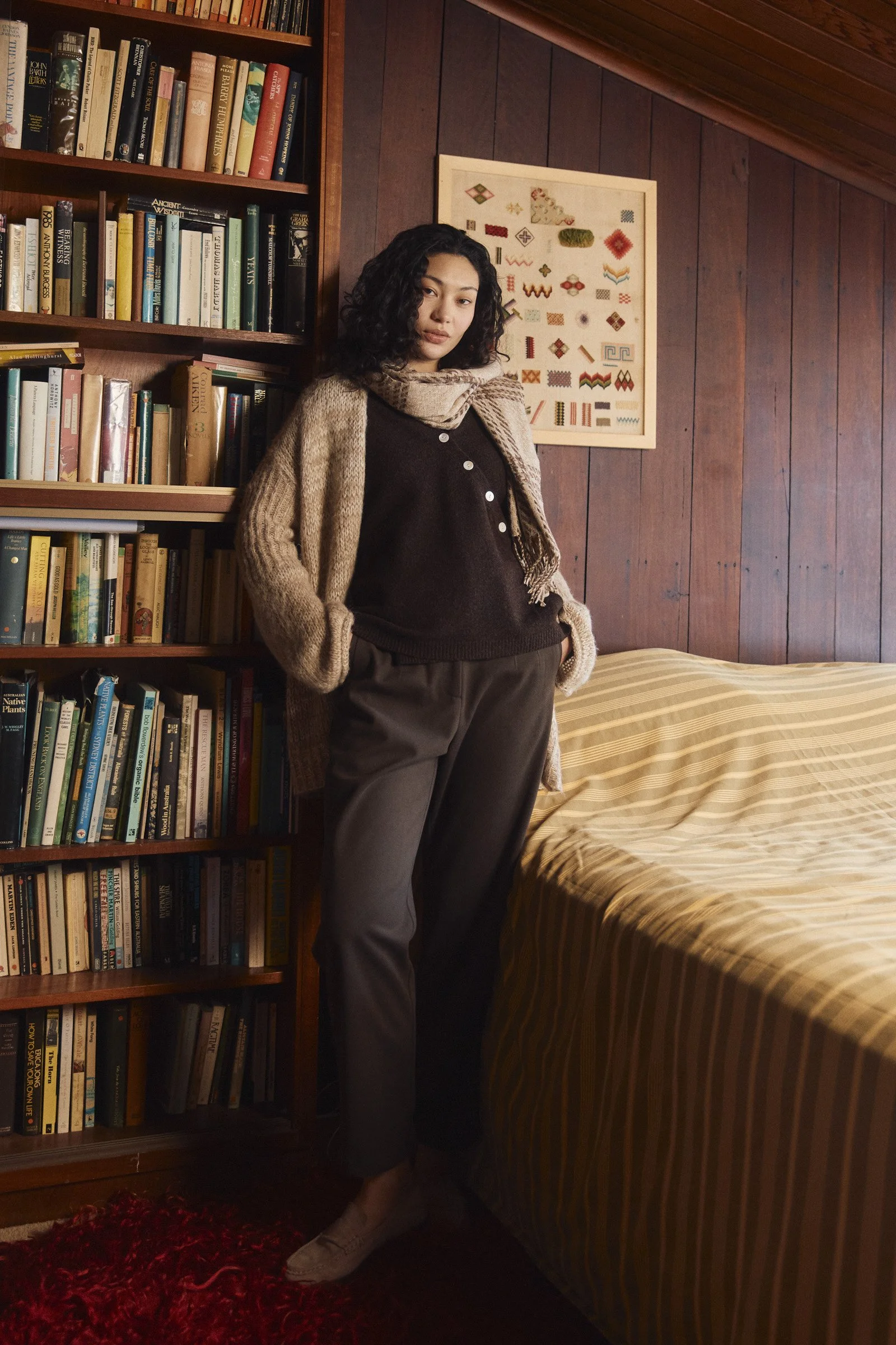 Woman with curly hair leaning against a wooden bookshelf filled with books, wearing a beige cardigan, black top, and gray pants, standing in a cozy, wood-paneled room.