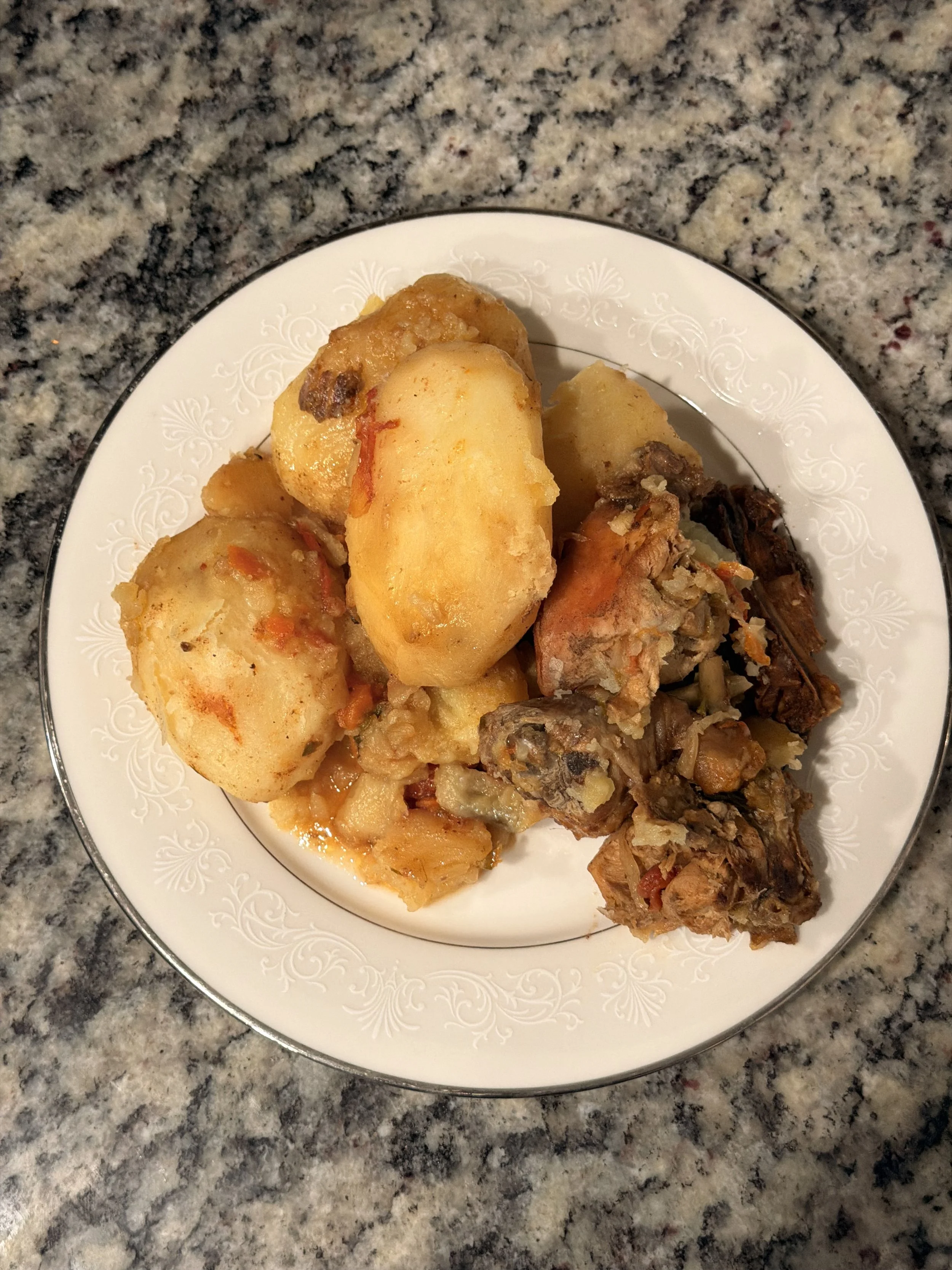 A white plate with a floral pattern on the rim holding several pieces of cooked potatoes and a piece of cooked meat, placed on a speckled granite countertop.