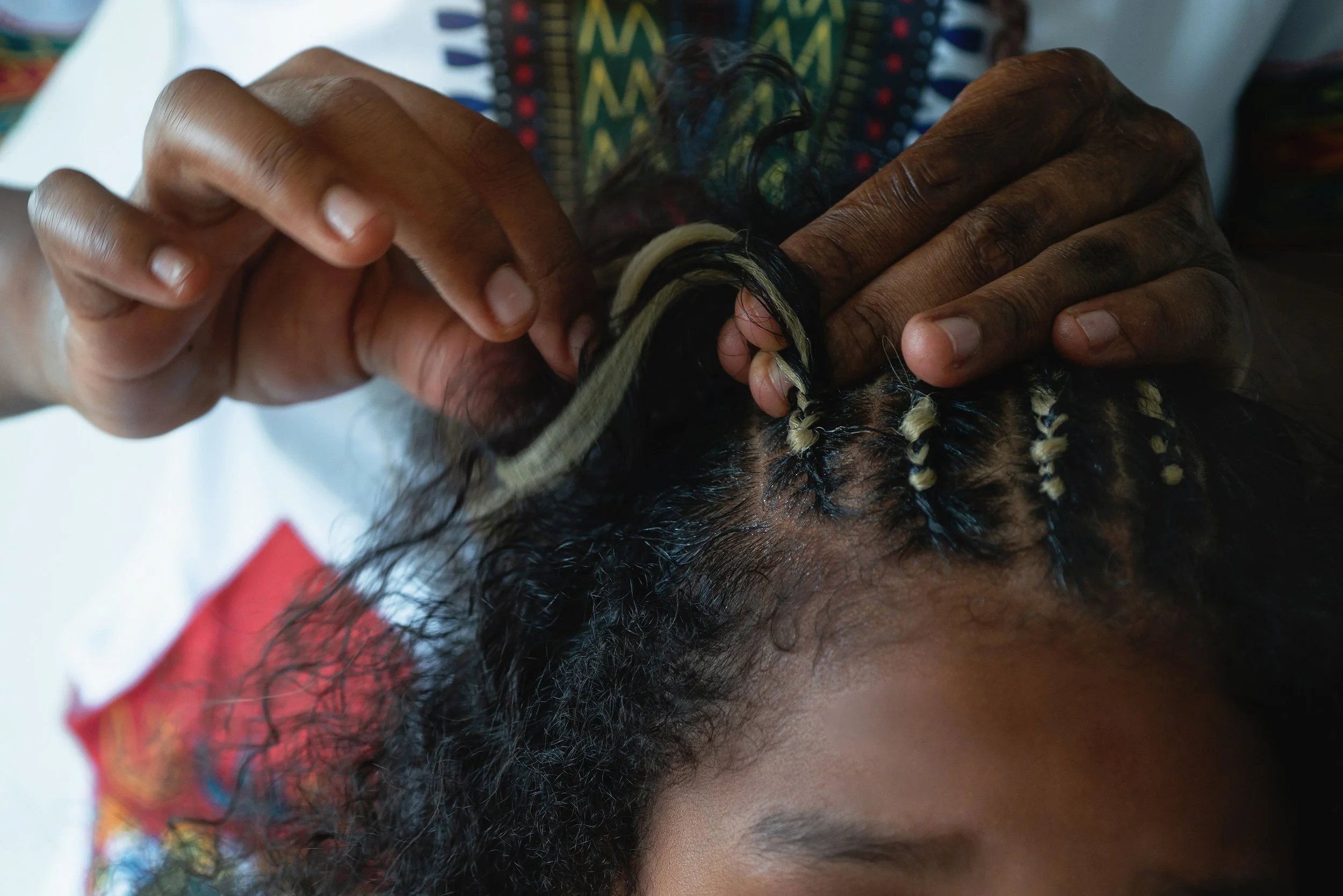 A hairstylist’s hands braiding intricate cornrows into a client’s hair, incorporating blonde extensions for a detailed and professional finish.