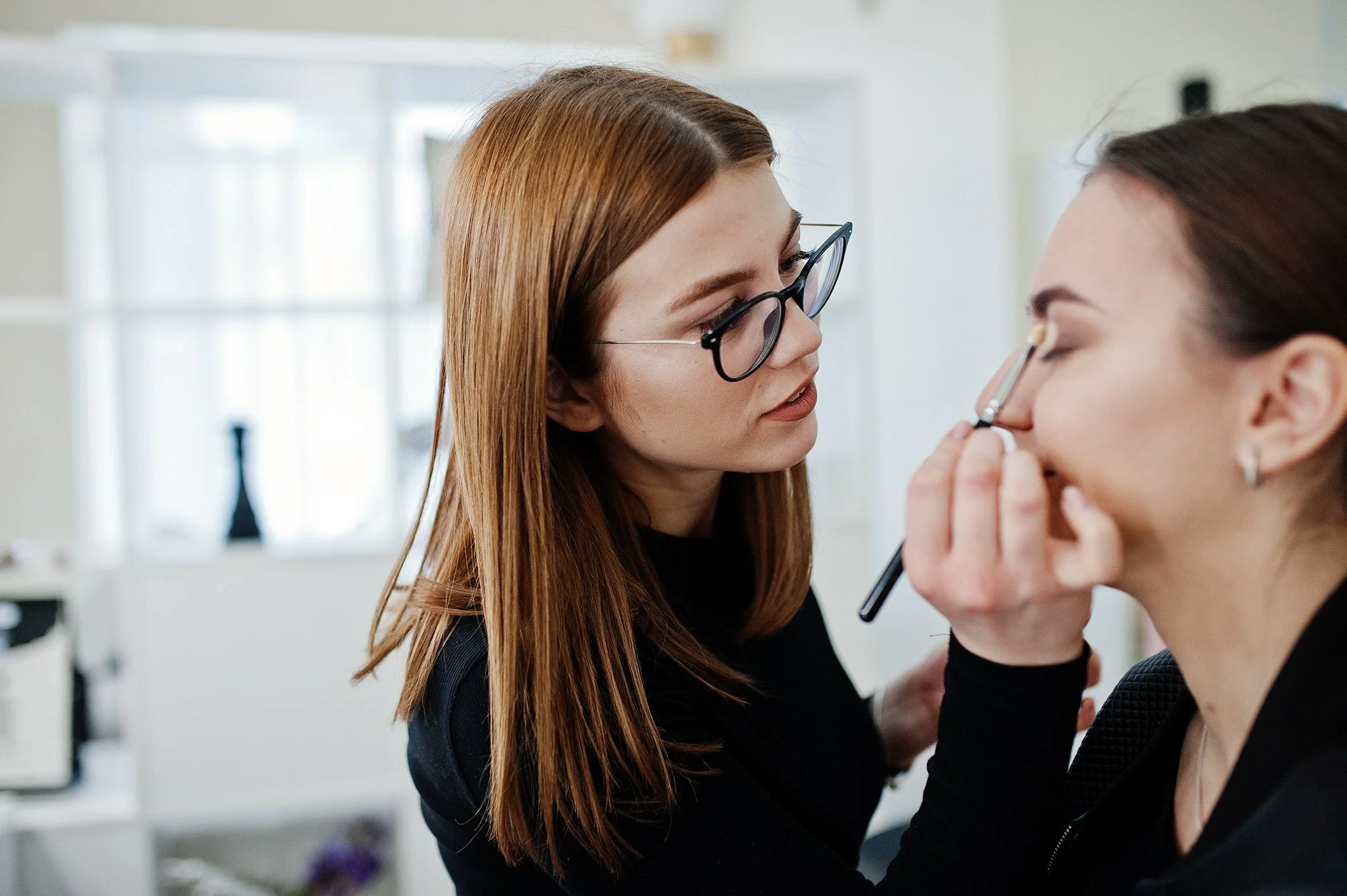 A makeup artist wearing glasses applying eyeshadow with a brush to a smiling client in a well-lit beauty studio.