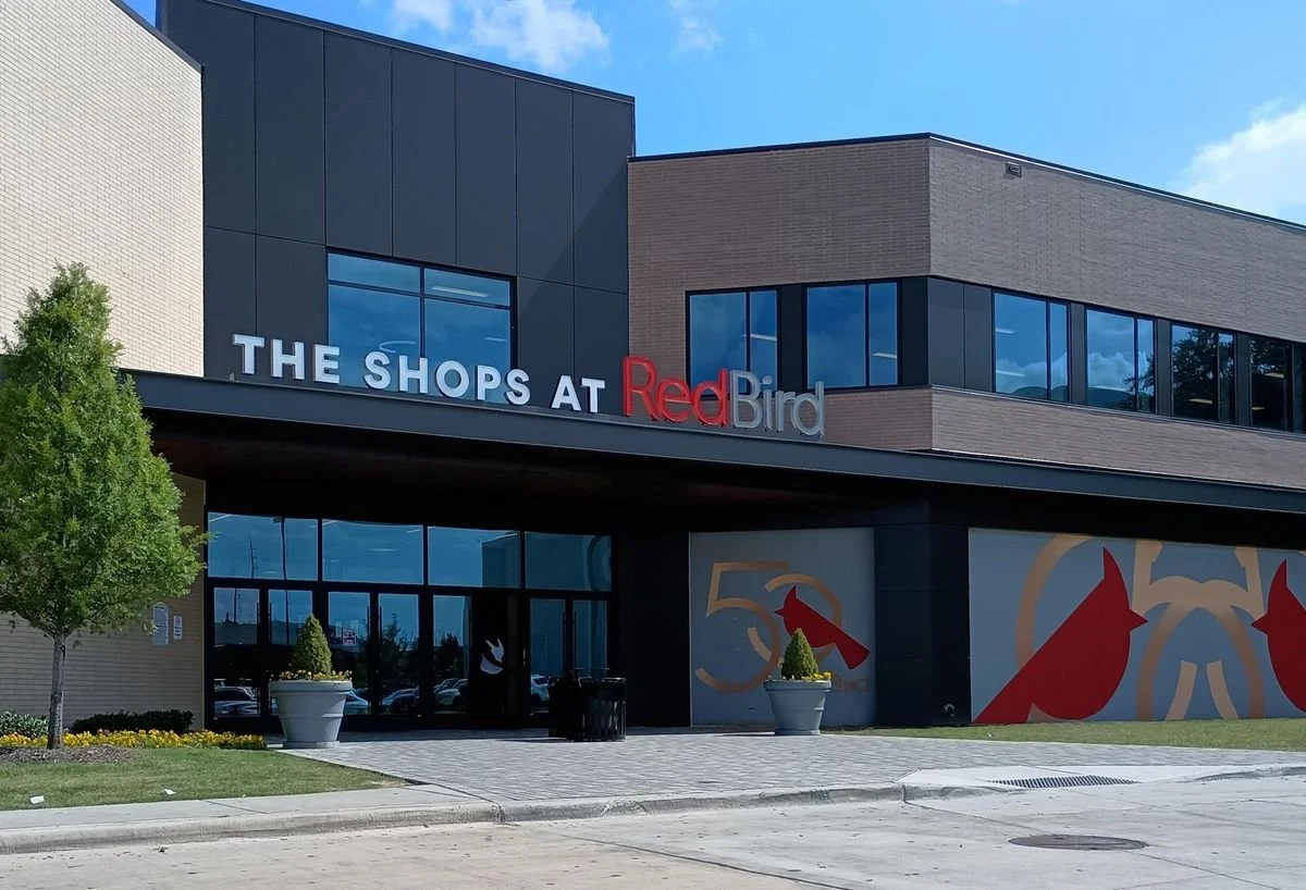 Exterior view of a shopping center called The Shops at Red Bird with modern architecture, large windows, and colorful signage.