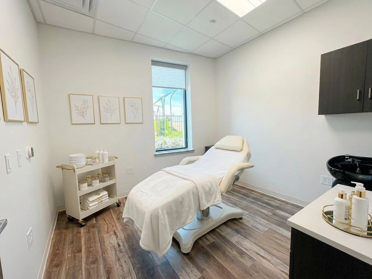 Medical treatment room with a white adjustable examination bed, a small white rolling cart with towels, bottles, and jars, a black sink with bottles on the counter, and tasteful wall decor of framed botanical prints. Window shows an outdoor playground.