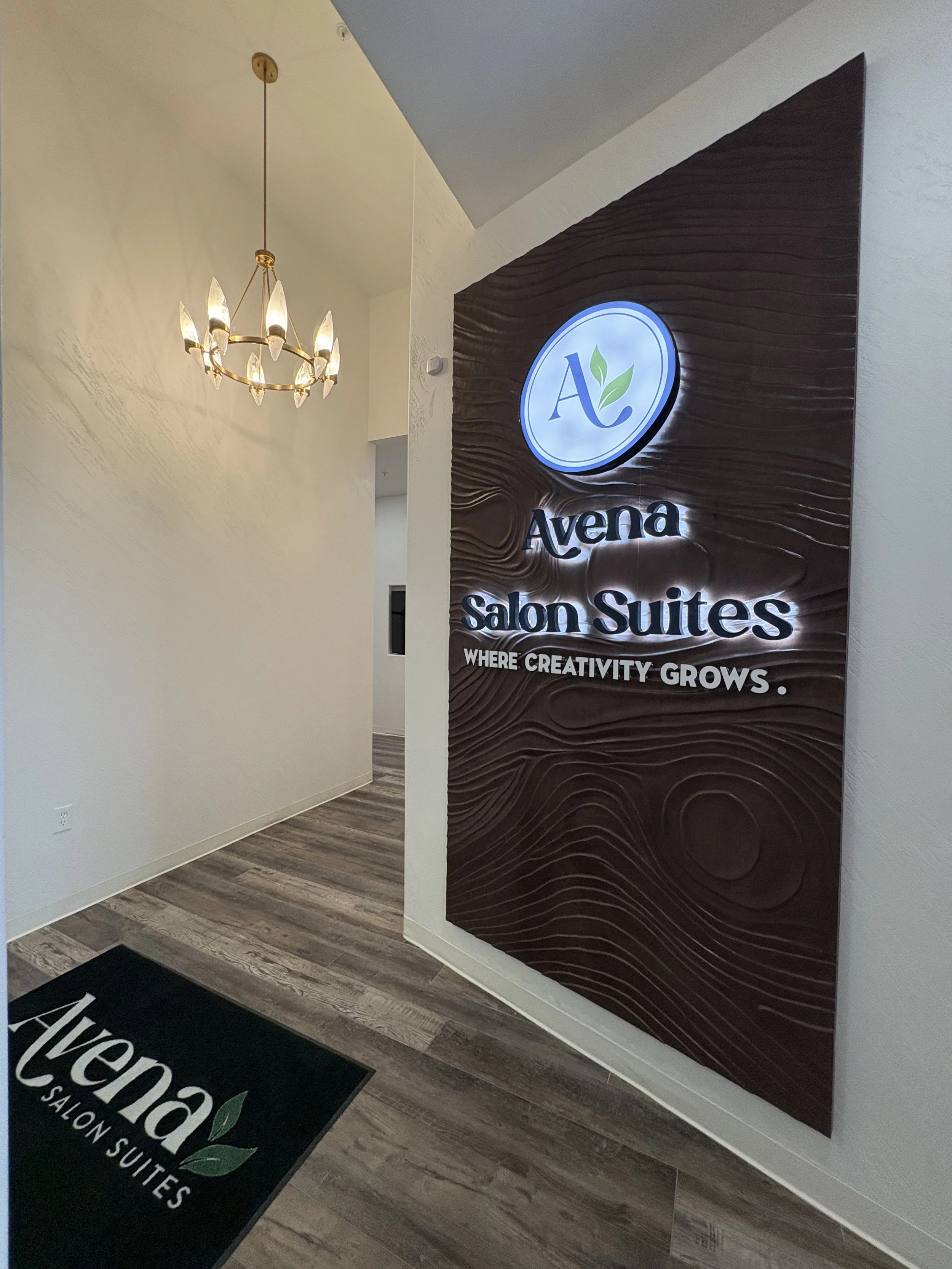 Interior view of a salon entrance with a chandelier, a sign reading 'Avena Salon Suites' on a textured wall, and a black mat with the same branding.