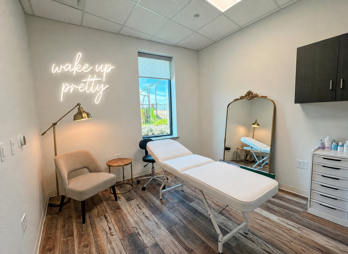 A medical or wellness treatment room with a massage or examination table, a chair, a small round table, a large mirror, a wall cabinet, and a neon sign reading 'wake up pretty'