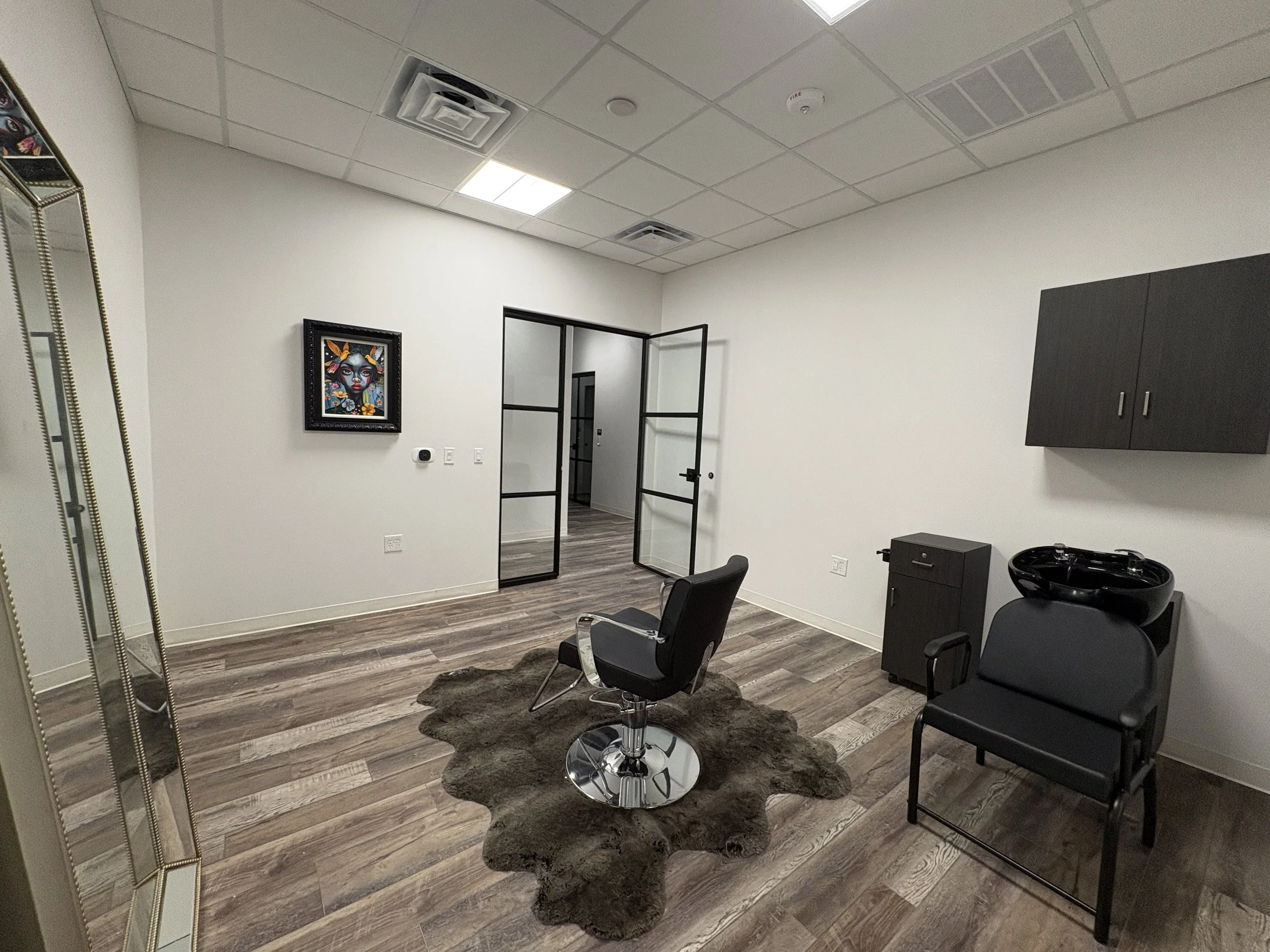 Interior of a modern salon or hair studio with black chairs, a dark cabinet, a decorative mirror, framed artwork, and a hair washing station with a black basin. The room has wood-style flooring and white walls.