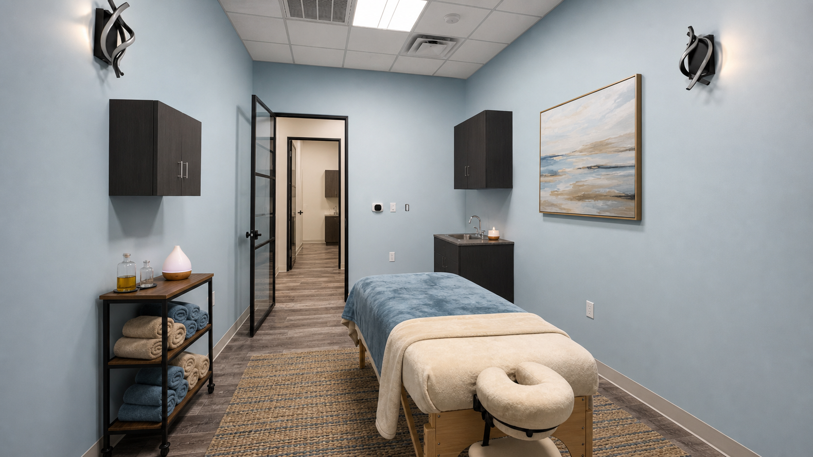 A therapy or massage room with a massage table covered with blue and beige towels, black cabinets on light blue walls, a small black cart with rolled towels and bottles, framed landscape painting, and a sink with a lit candle.