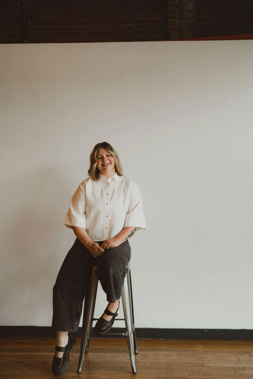 A woman sitting on a tall metal stool against a plain white wall, smiling, wearing a cream-colored blouse, black pants, and black shoes, with a tattoo on her left forearm.