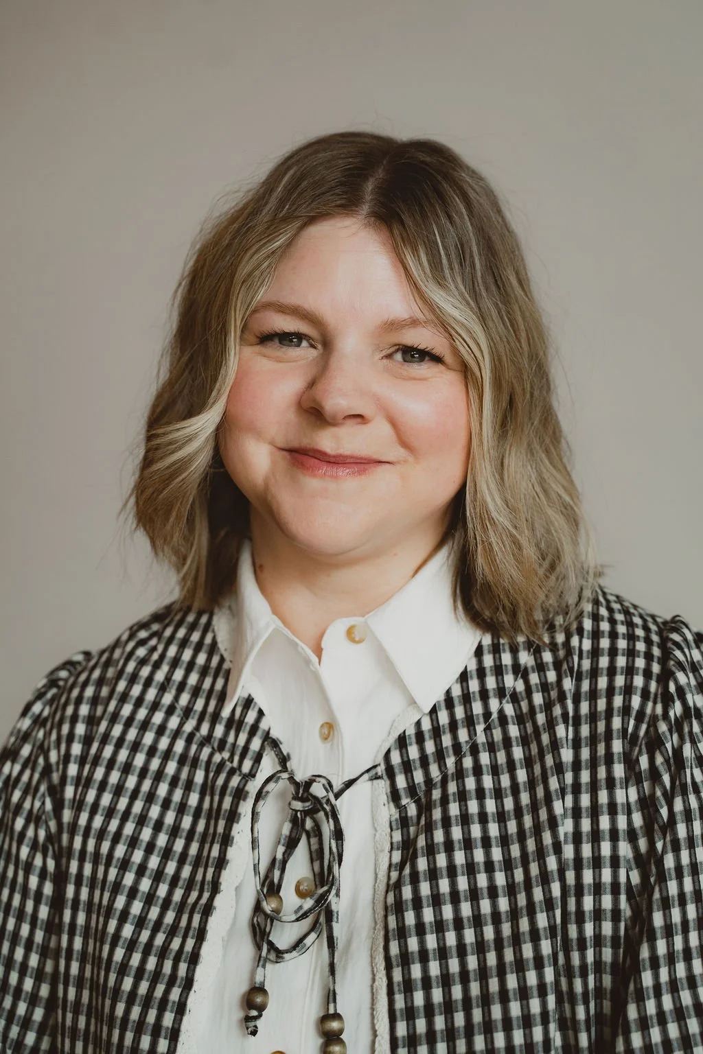 A woman with shoulder-length wavy blonde hair, wearing a white collared shirt and a black and white checkered jacket, smiling softly against a plain background.