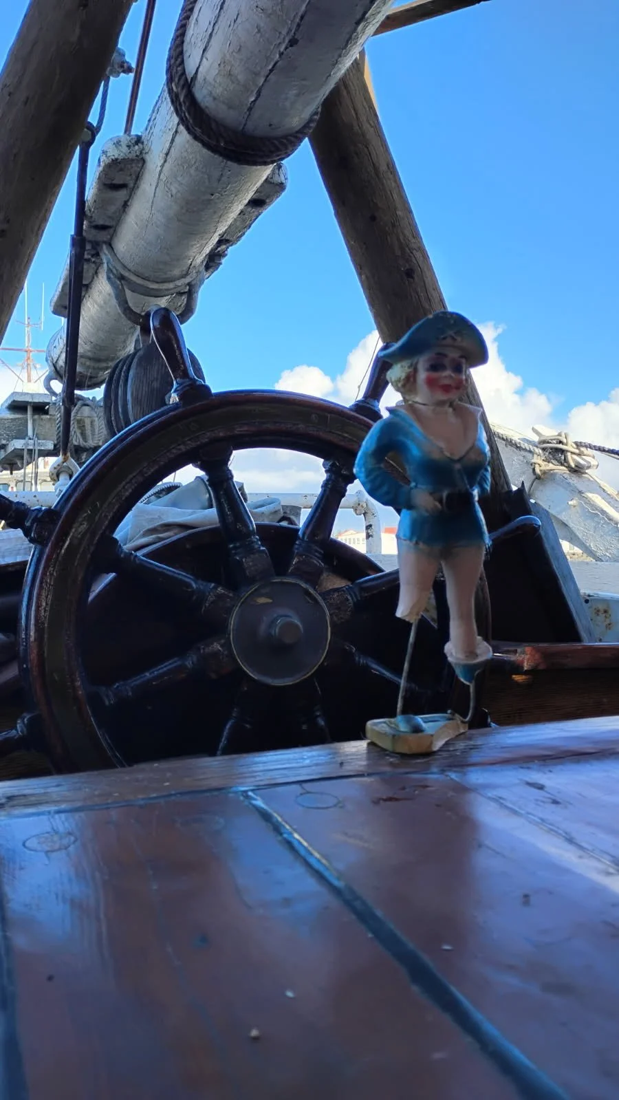 Close-up of a ship's steering wheel with a pirate figurine in a blue coat and black hat standing nearby, against a background of a blue sky with some clouds.