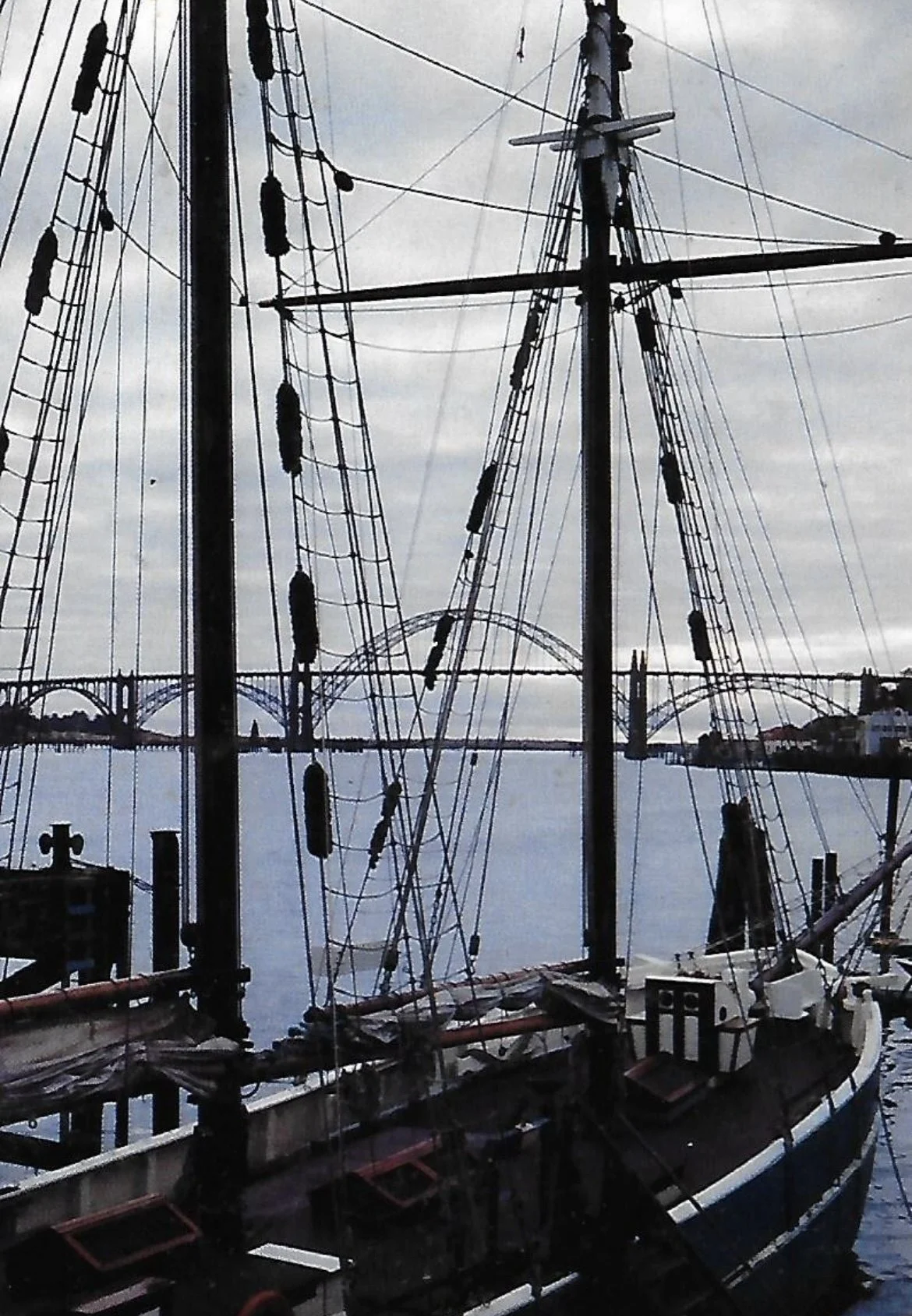 View of a large sailing ship's masts and rigging with a bridge and water in the background, overcast sky.
