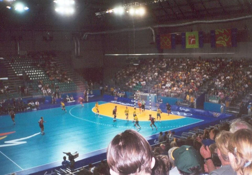 Handball game underway on a turquoise indoor court with players, spectators, and bright arena lights