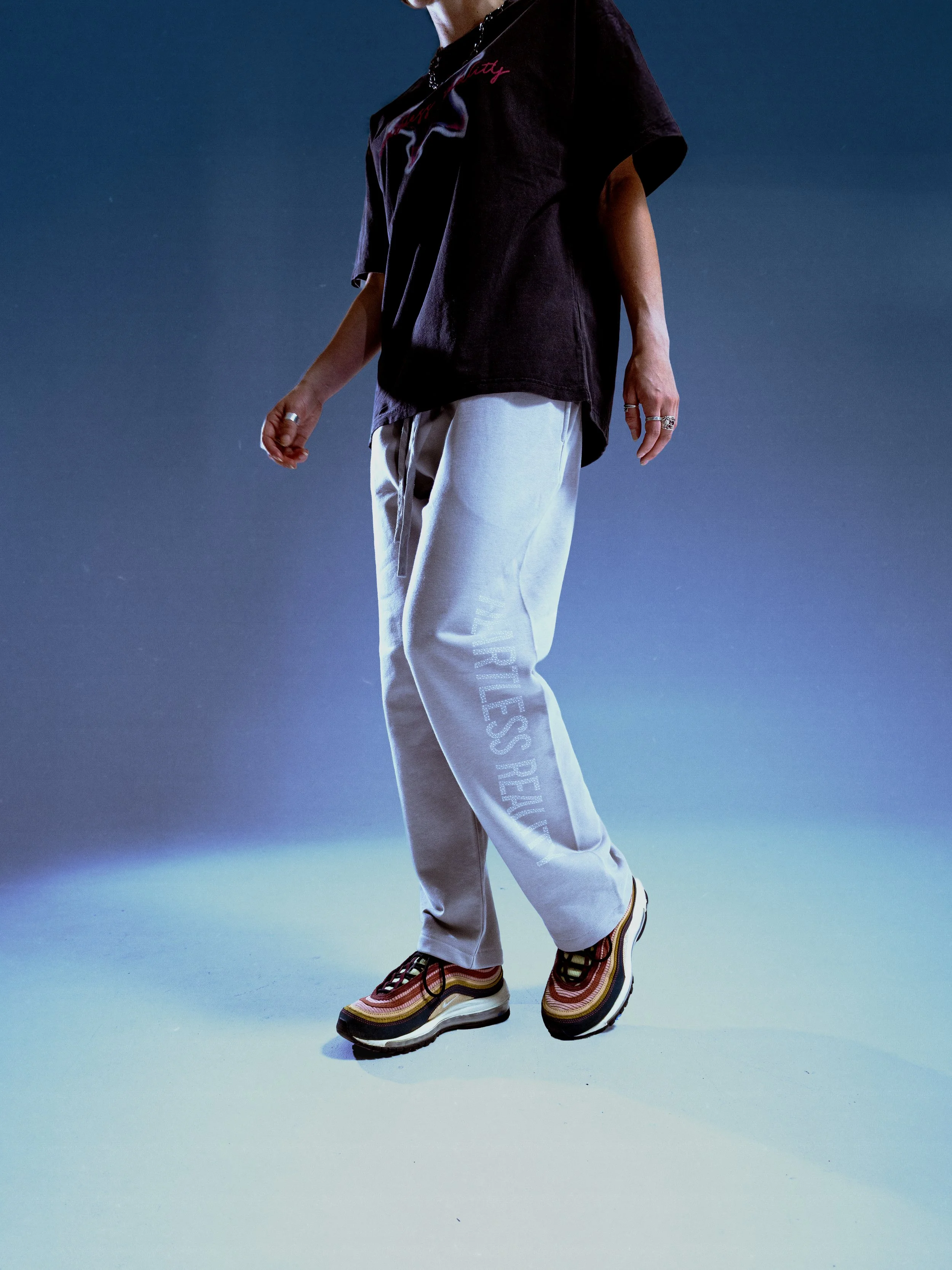 Close-up of a person wearing colorful striped sneakers, light gray sweatpants with white text on the left leg, a loose black T-shirt, and multiple rings on their fingers, standing against a gradient blue and green background.