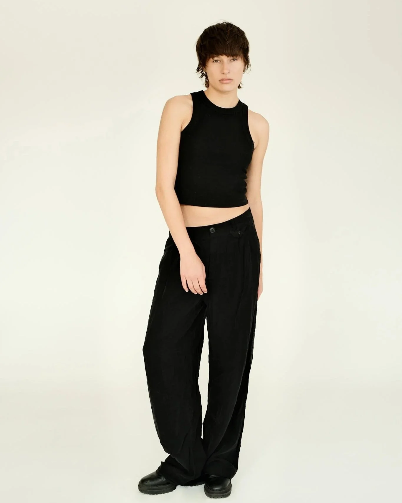 A person with short dark hair wearing a black sleeveless top and black baggy pants standing against a plain light background.