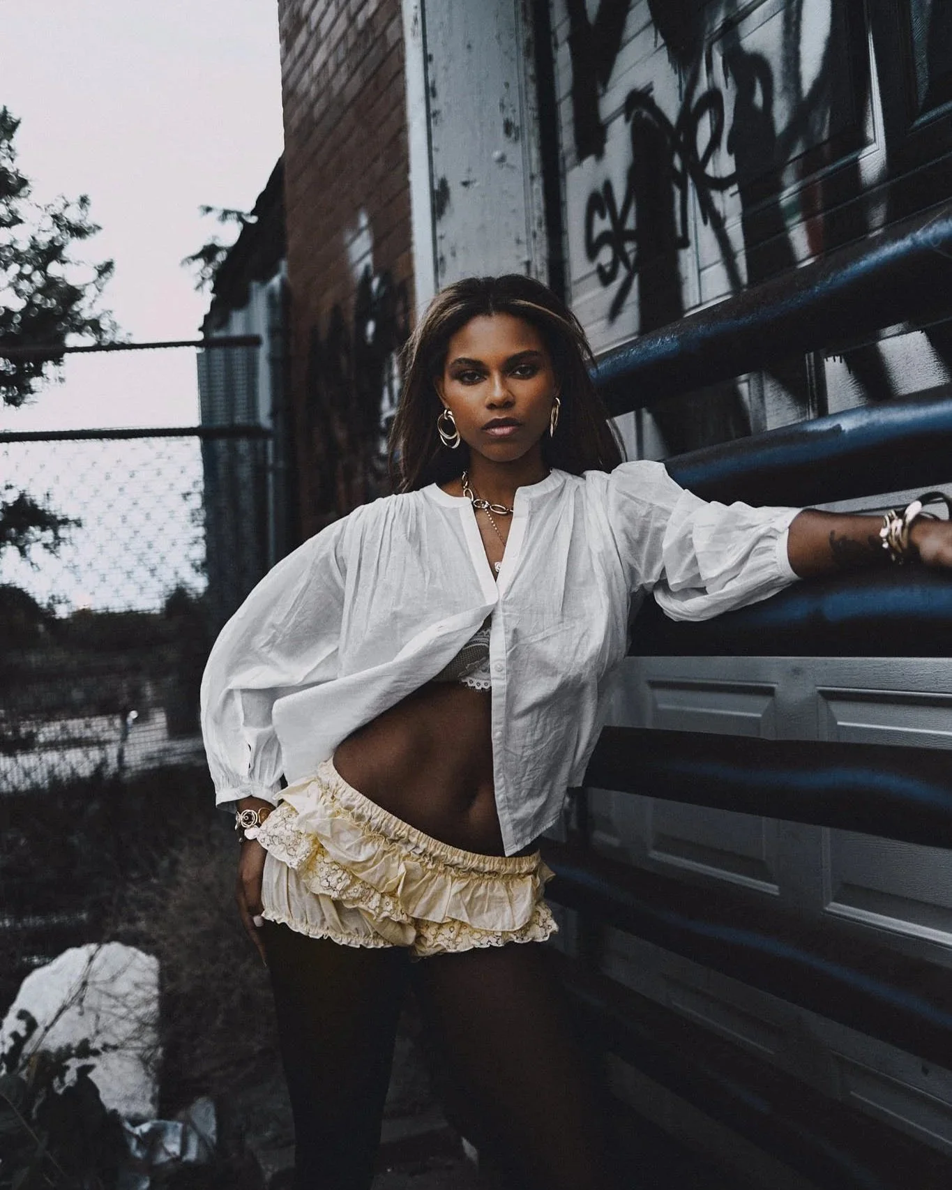 A woman with dark hair and dark eyes standing outdoors near a building with graffiti, dressed in a white long-sleeve blouse and a layered yellow skirt, accessorized with hoop earrings and jewelry.