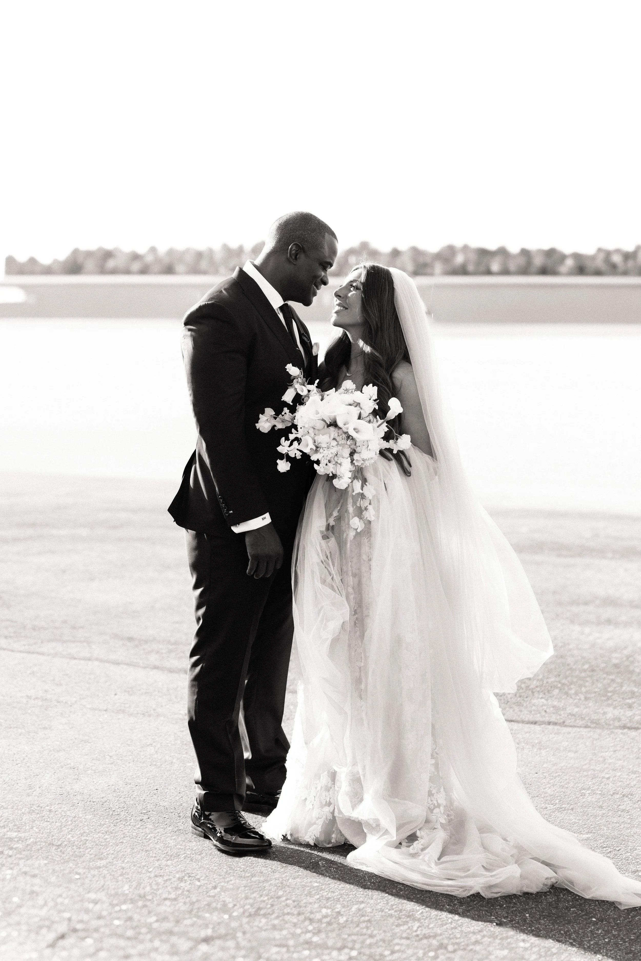 Black-and-white outdoor wedding portrait of a bride and groom smiling together on the airport tarmac with a bridal bouquet.