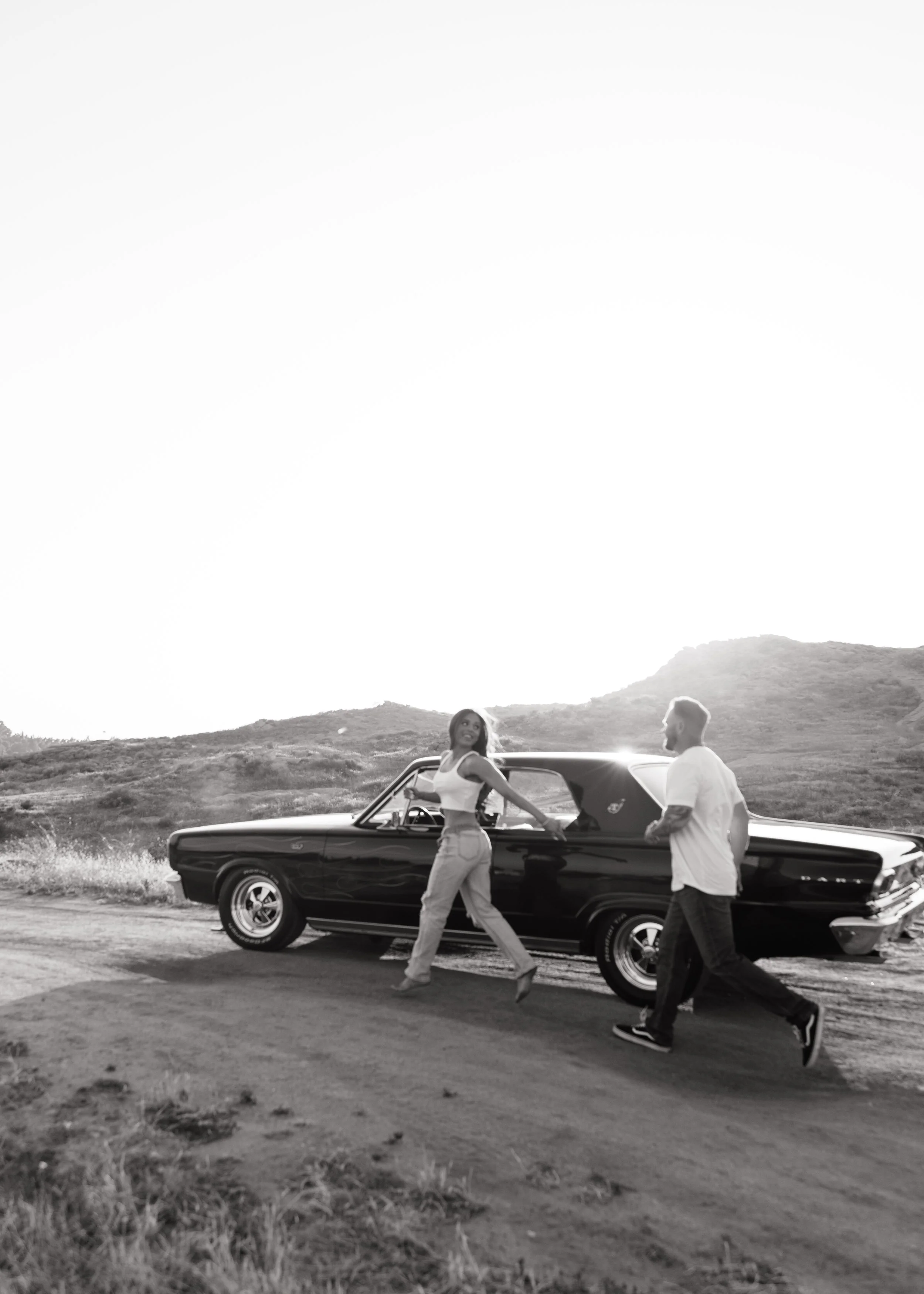 Black-and-white film-style photo of a couple walking beside a vintage convertible in an open outdoor landscape.