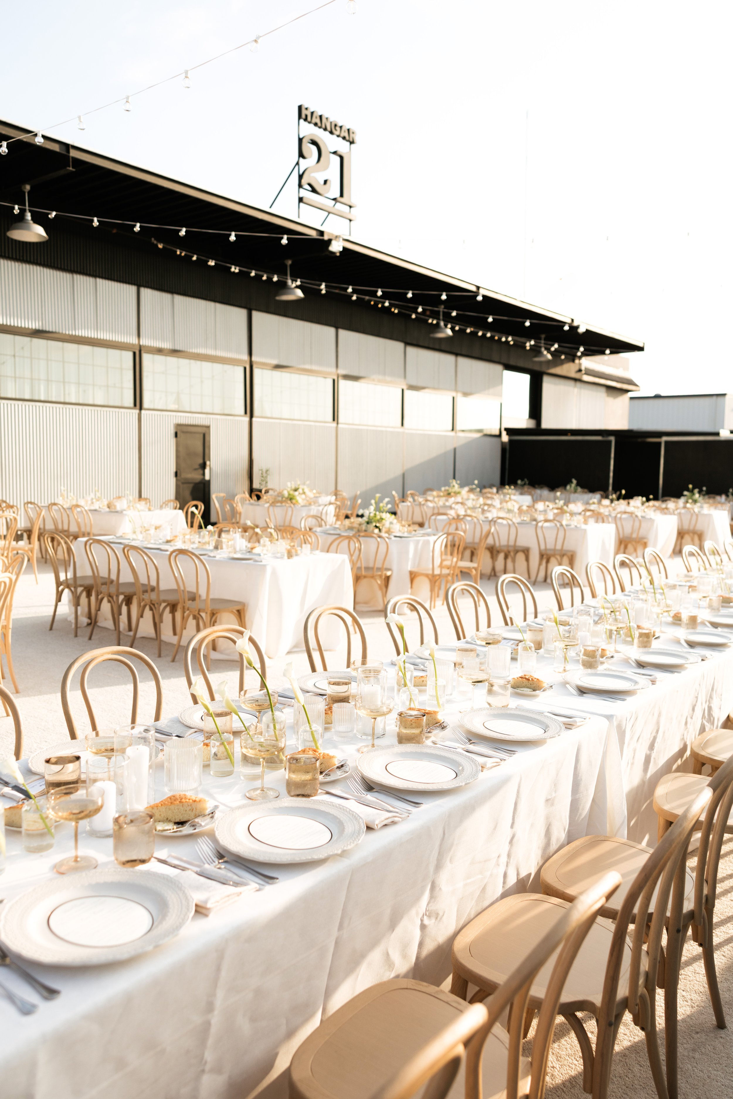 Outdoor wedding reception setup with long tables, white linens, wooden chairs, place settings, and string lights outside an industrial venue.