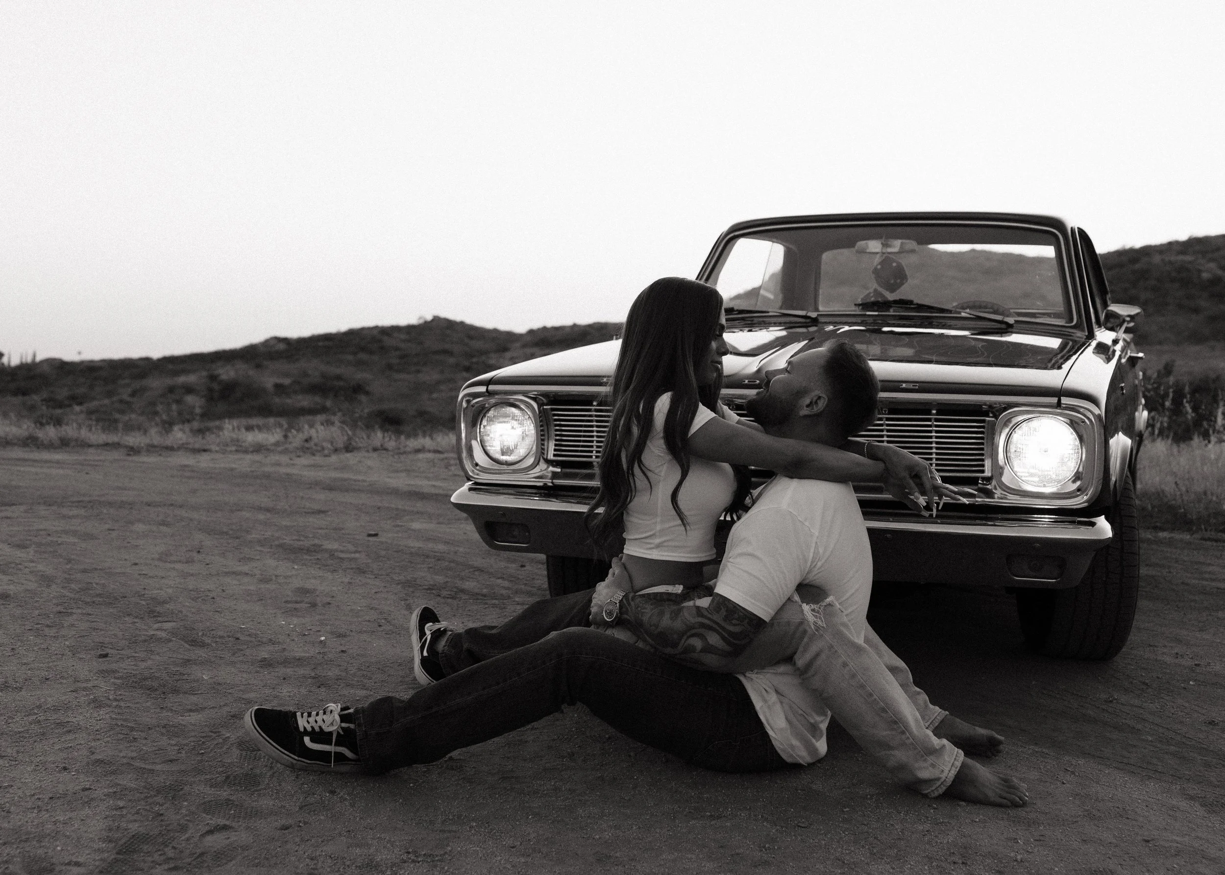 A man and woman sitting on the ground in front of a vintage car on an empty road with hills in the background, embracing each other.
