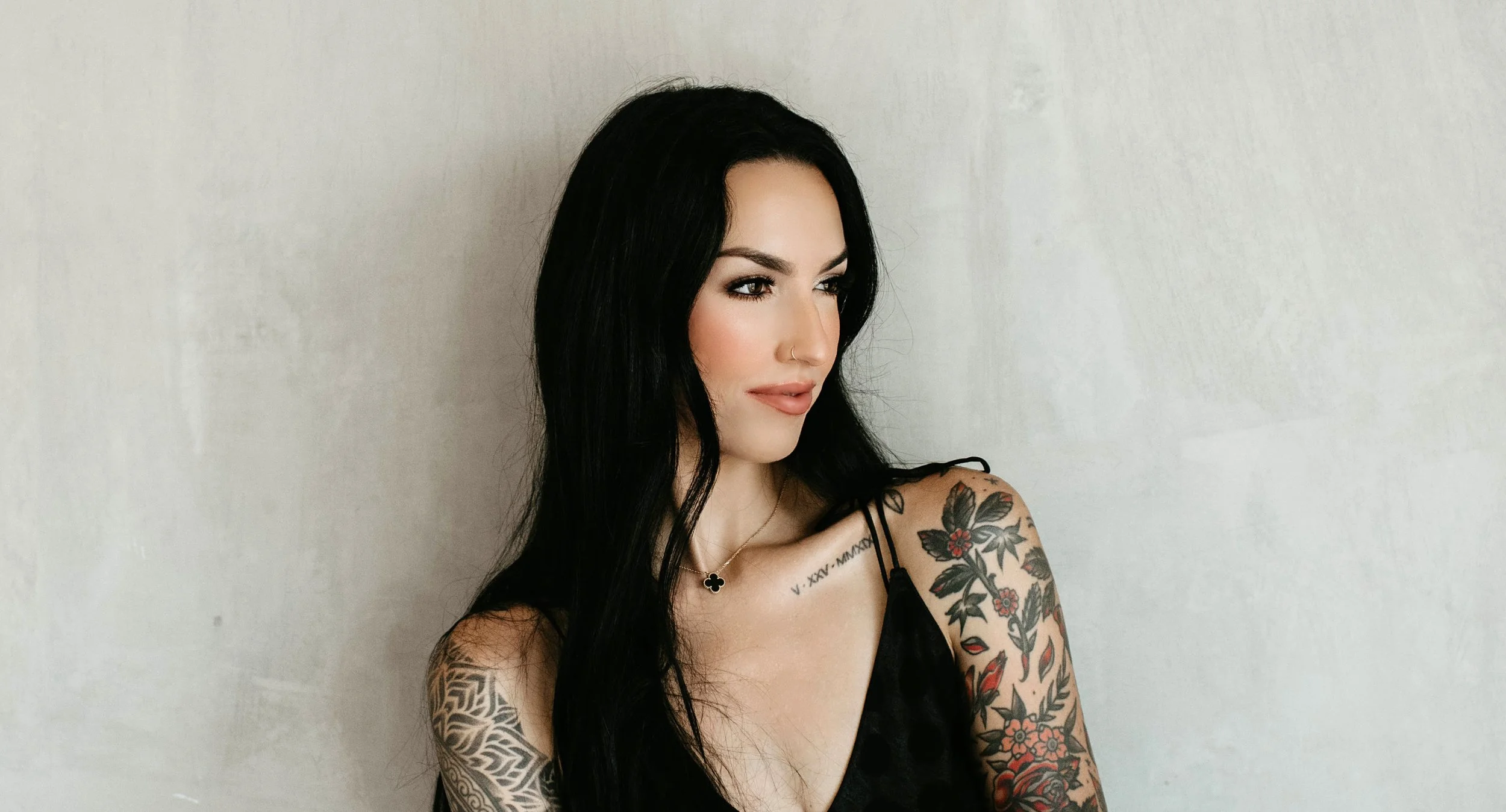 Ash Modiano - A woman with black hair, tattoos on her arms and shoulder, wearing a black top, standing against a plain white wall.
