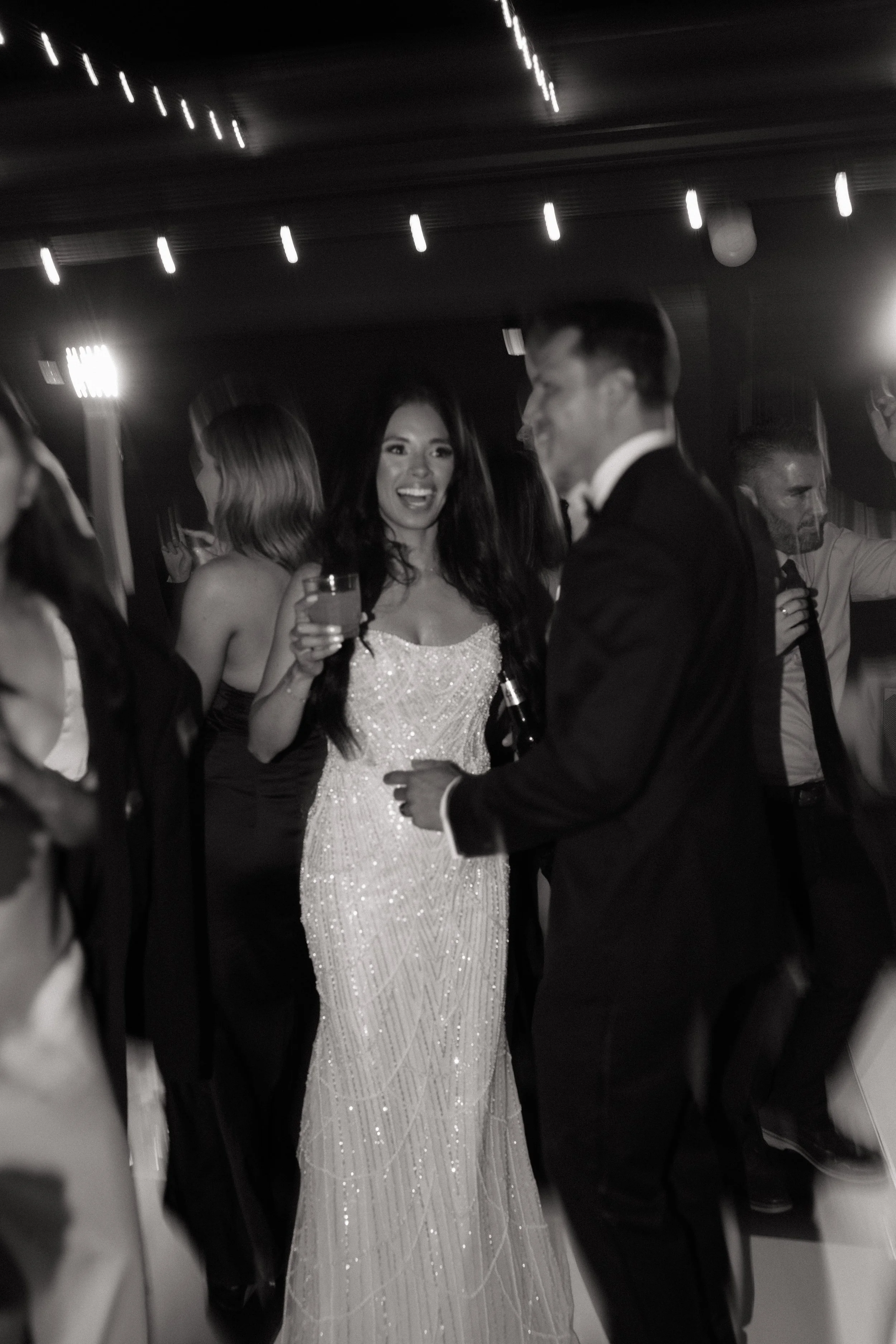 Black-and-white candid wedding reception moment with guests mingling under string lights at an outdoor venue.