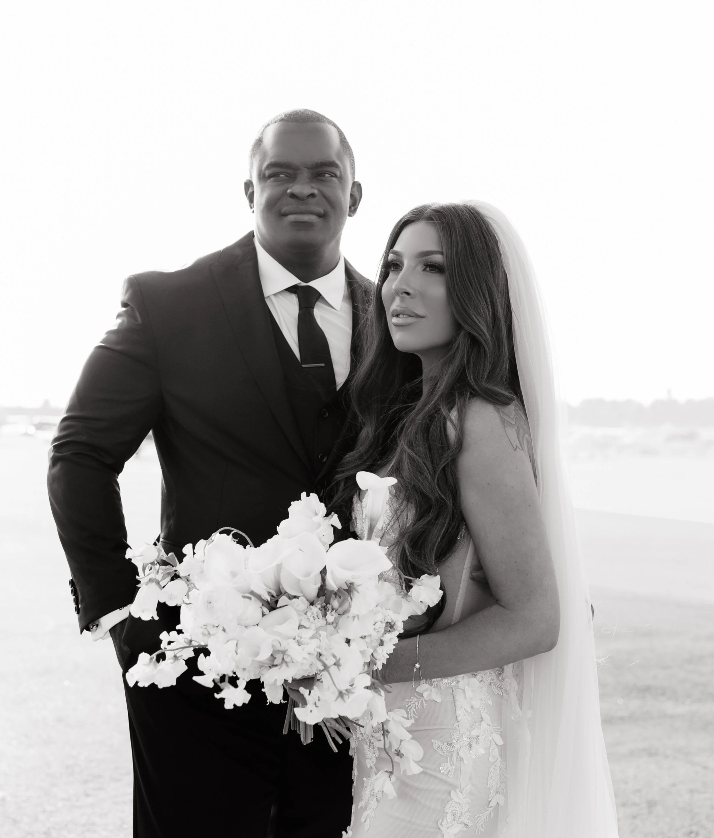 A black and white photo of a bride and groom outdoors, the groom wearing a suit and tie, and the bride holding a large bouquet of white flowers.