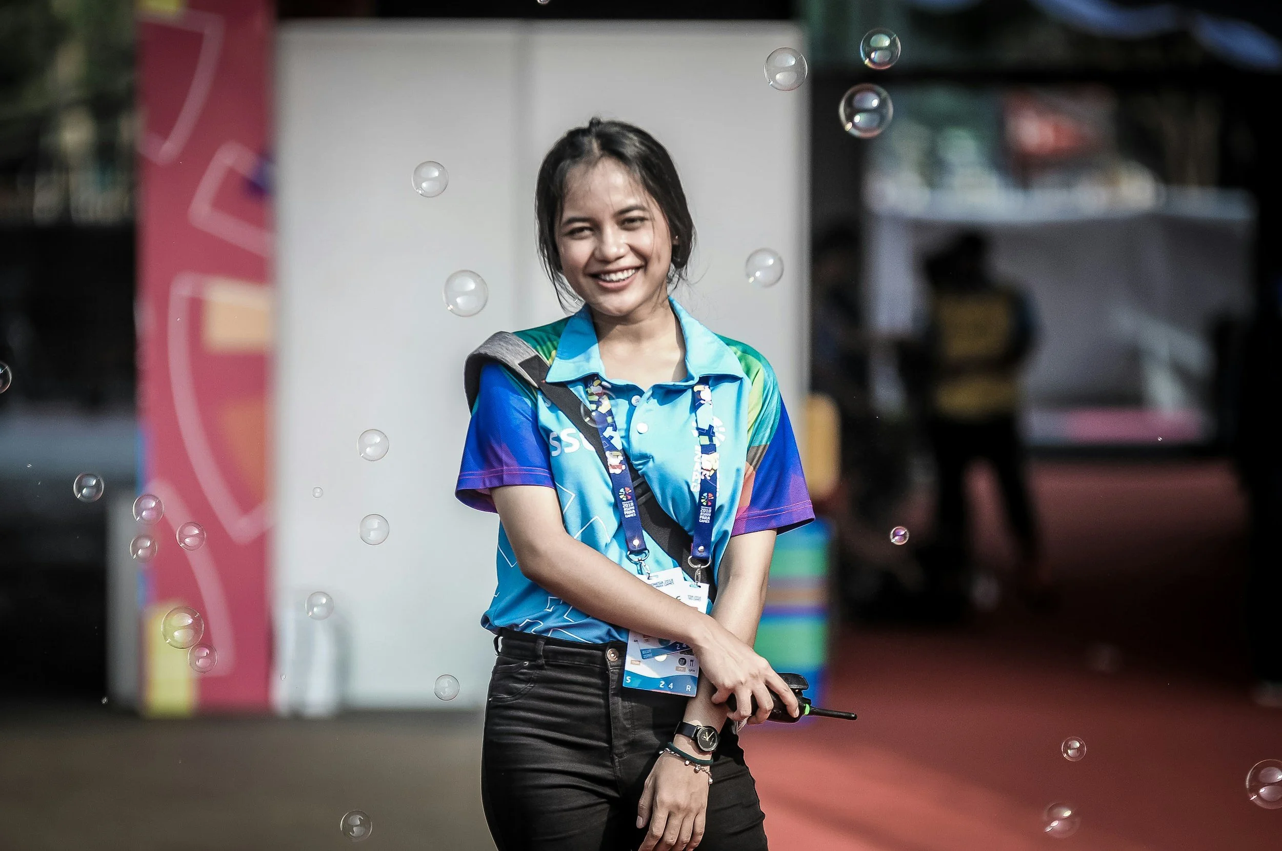 A young woman with dark hair and light skin, smiling, stands in an outdoor setting with bubbles floating around her. She wears a colorful sports shirt and has an ID badge hanging around her neck. The background is blurred with various objects and colors.