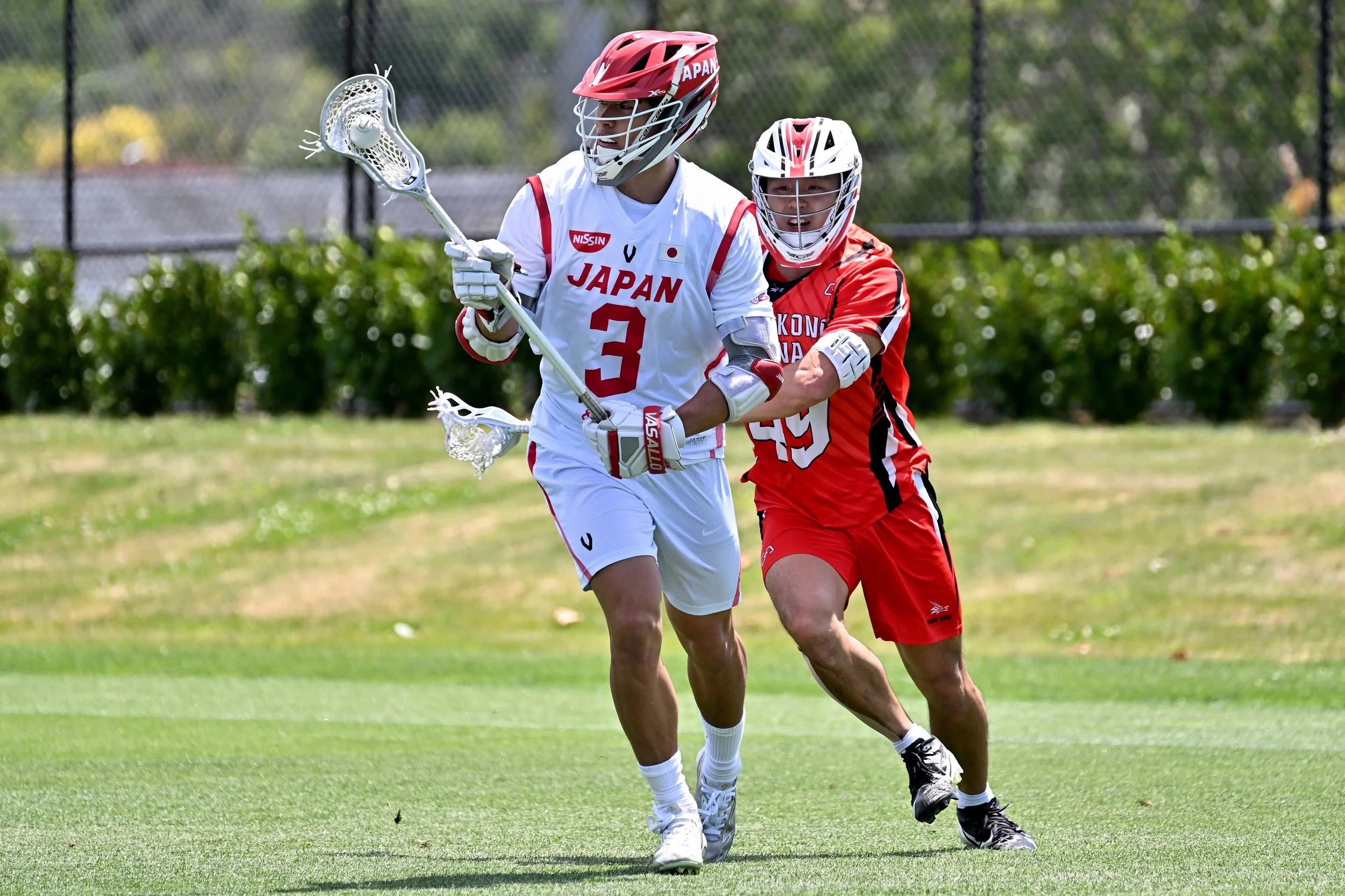 Two athletes playing lacrosse, wearing helmets and uniforms representing Japan, on a grassy field with trees and a fence in the background.