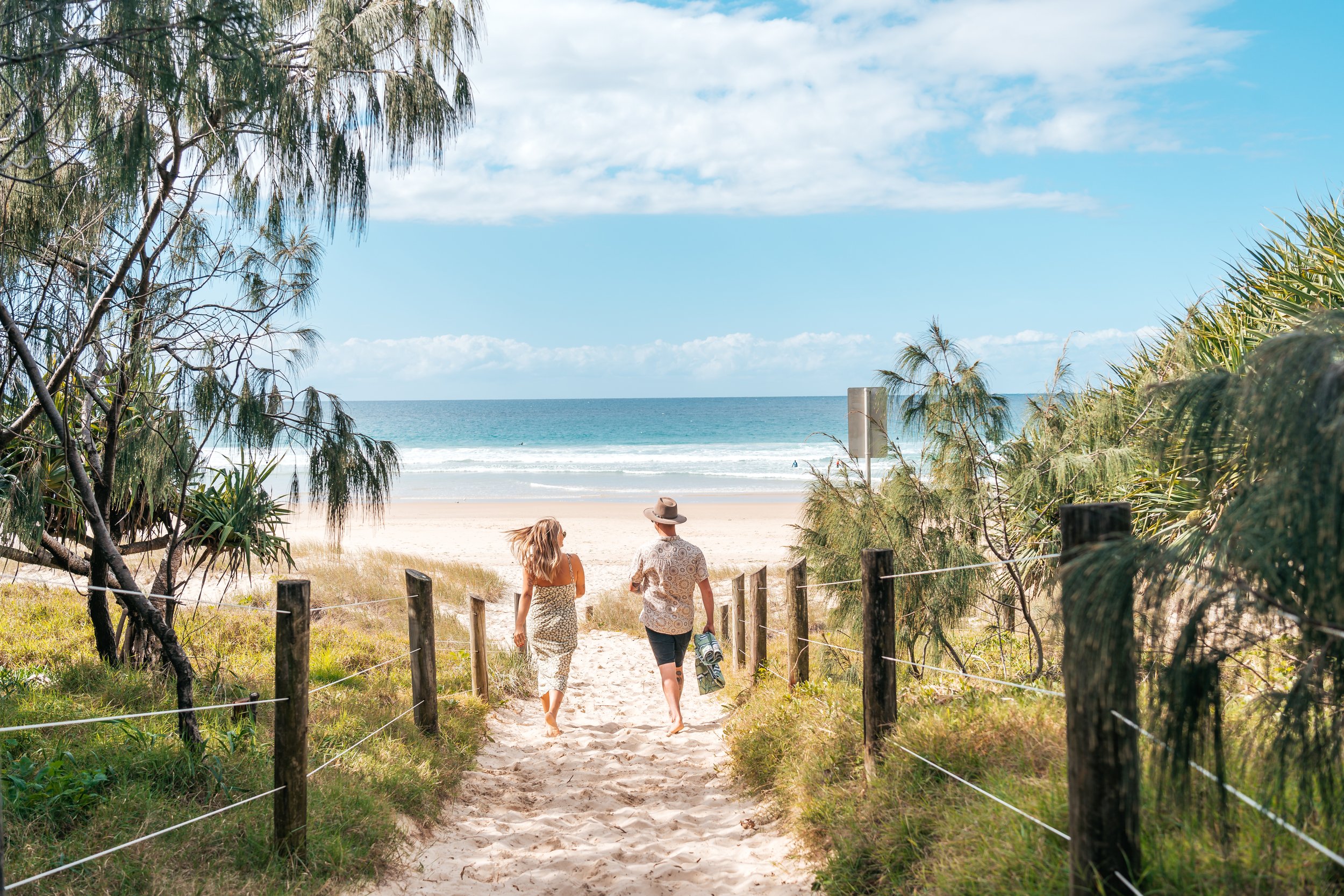 A man and woman walking down a sandy beach path towards the ocean, framed by trees and a wooden fence, on a sunny day with blue sky and scattered clouds.